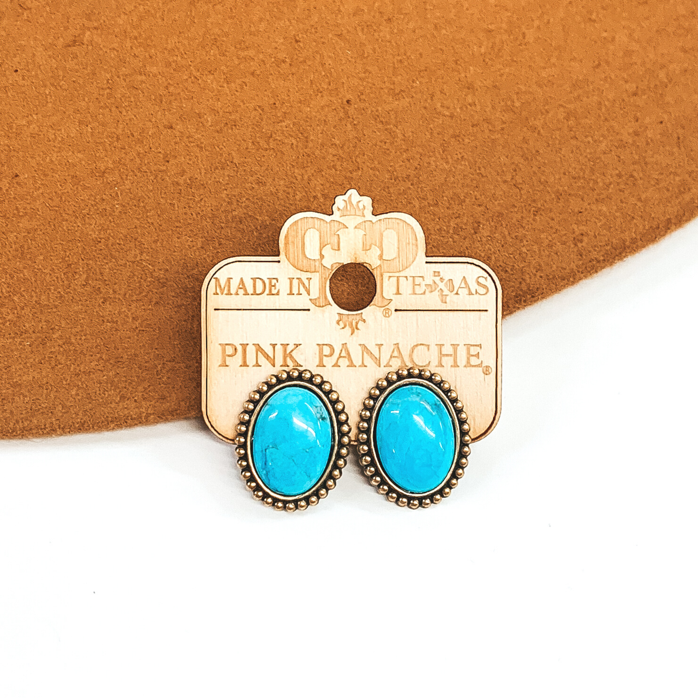 Bronze set earrings with tiny bronze beads surrounding the outside of the earrings. There is a center oval turquoise stone. These earrings are on a camel colored and white background. 