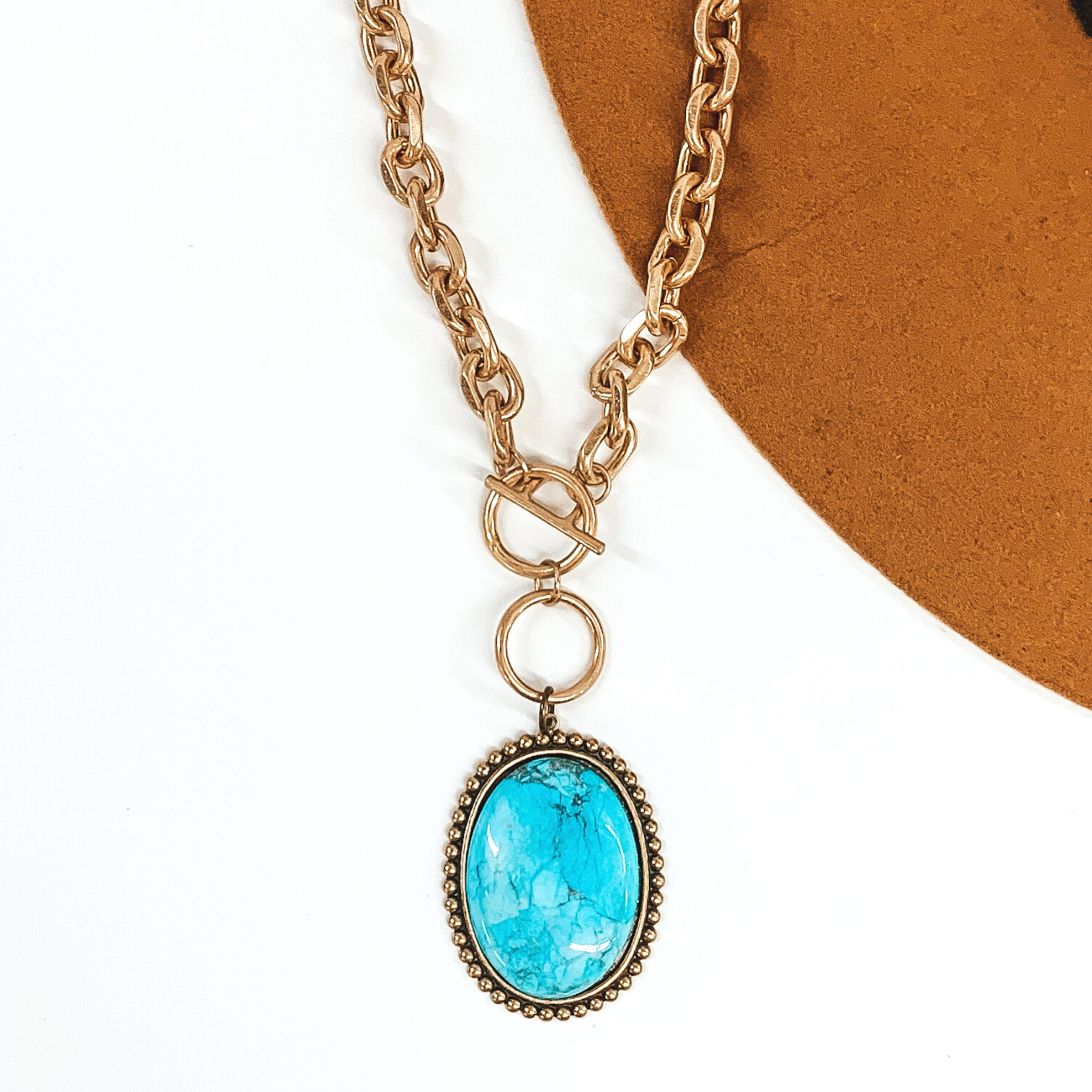 Bronze set oval pendant with turquoise cabochon stone. It is hanging from a toggle front clasp on a matte gold chain link necklace. This necklace is pictured on a white and camel background. 