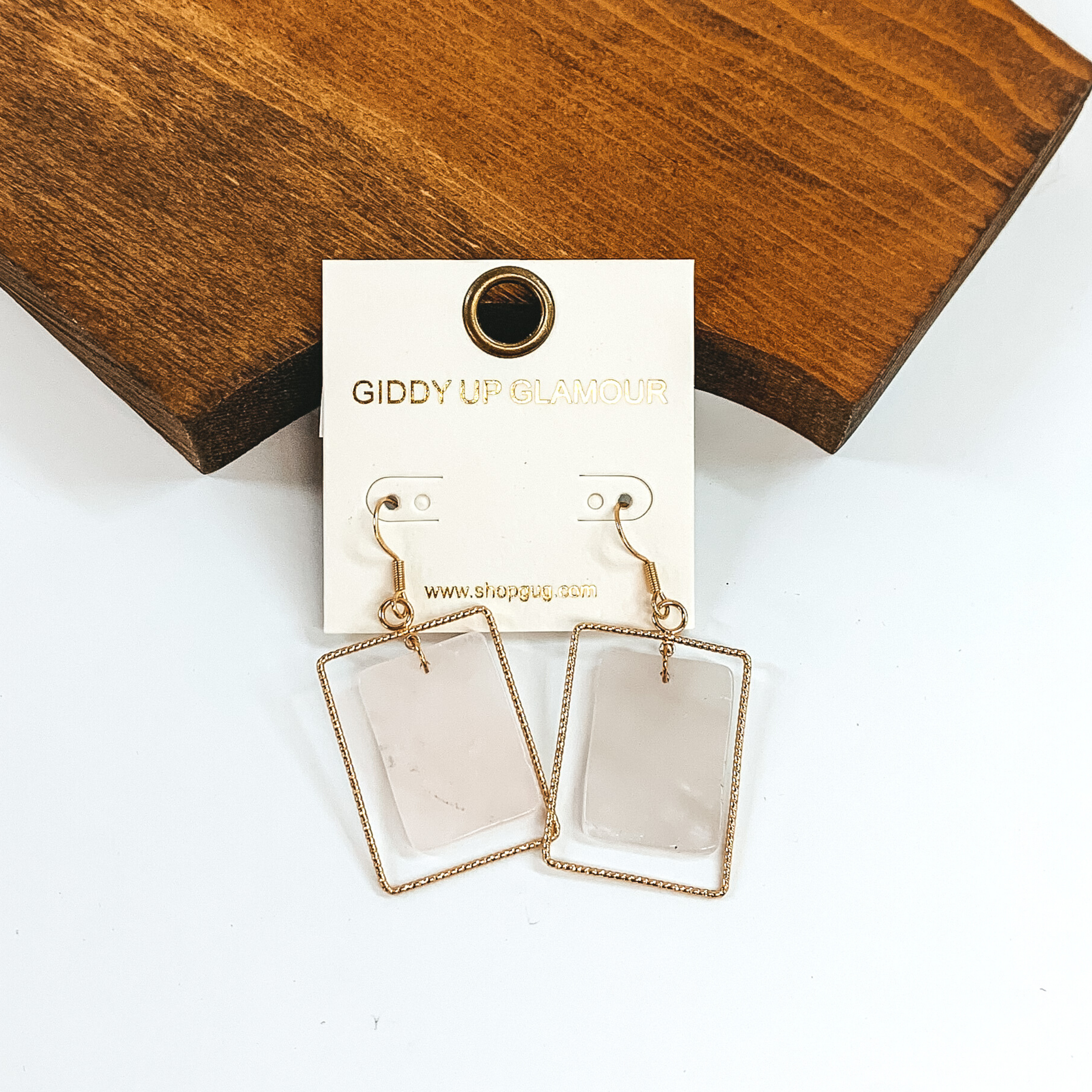 These earrings have a thin gold dangle rectangle with a center rectangle rose colored stone. These earrings are pictured laying on a dark wood piece on a white background. 