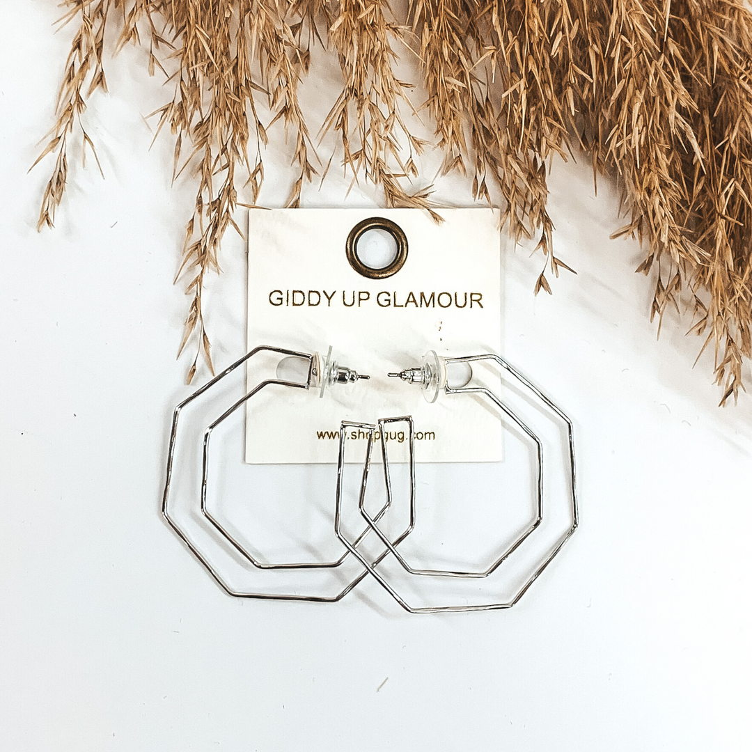 Thin, silver hexagon shaped hoop earrings. They are pictured on a white background with tan floral at the top of the picture. 