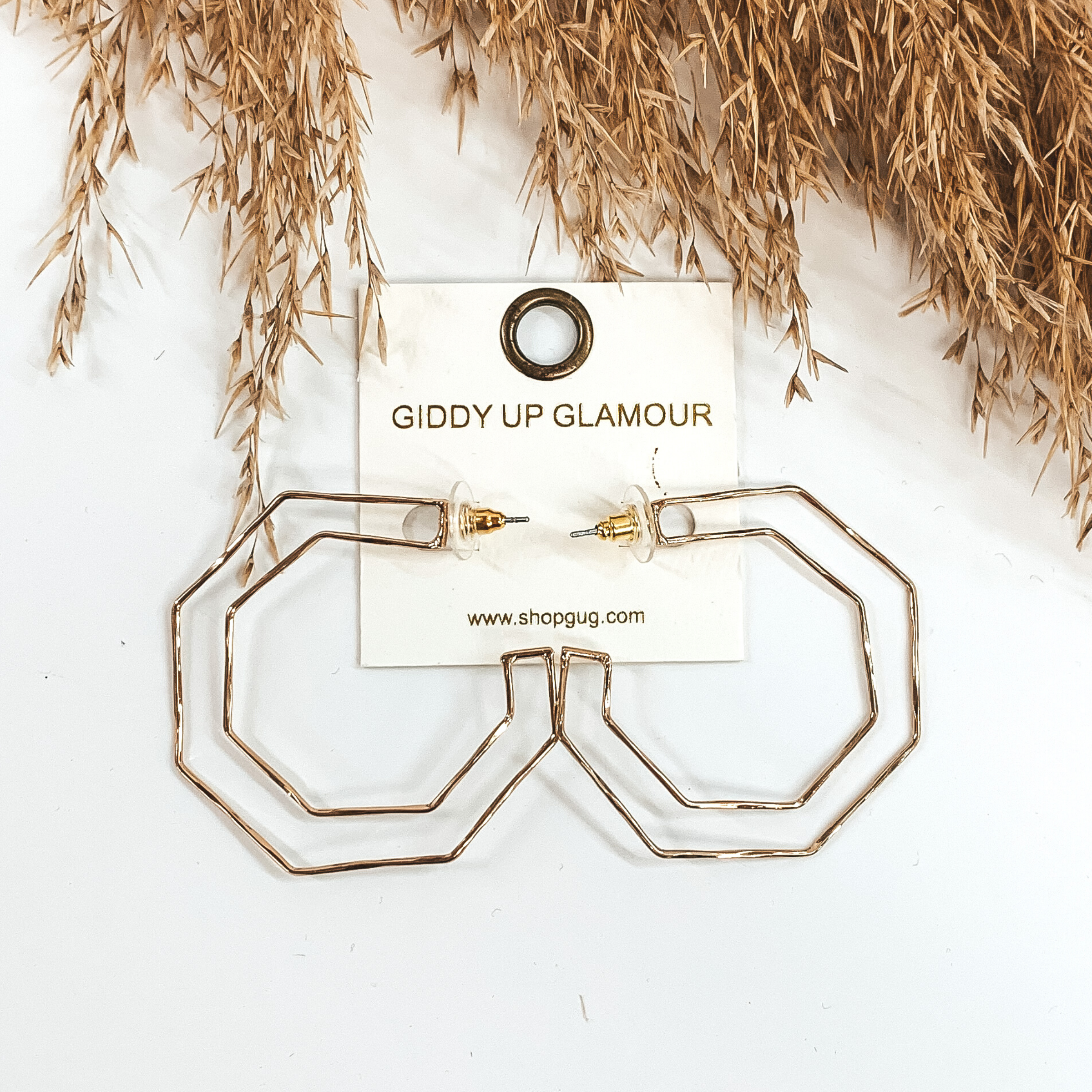 Thin, silver hexagon shaped hoop earrings. They are pictured on a white background with tan floral at the top of the picture.