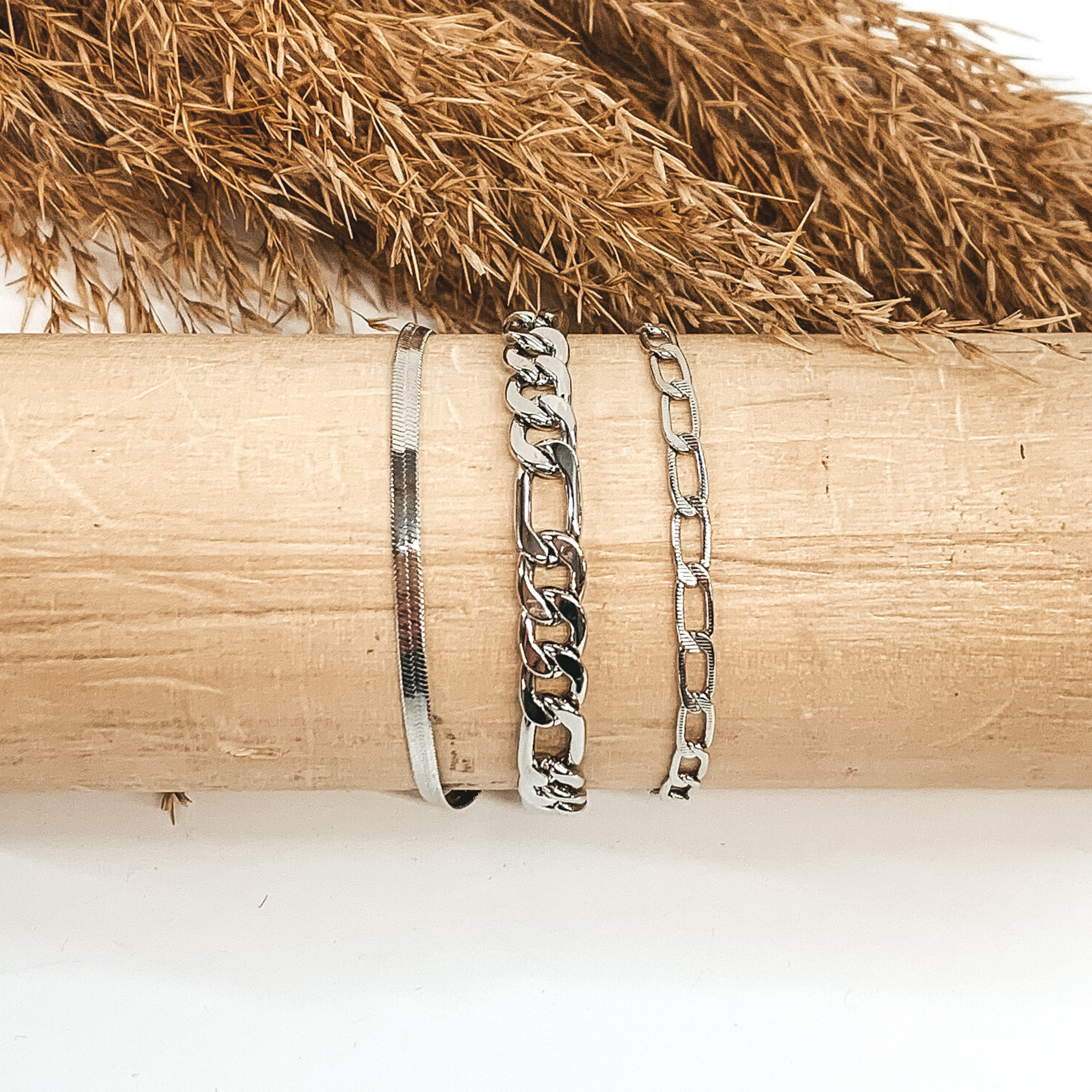 This silver bracelet includes three different types of chains. This bracelet is on a light colored piece of wood pictured on a white background with some tan floral at the top of the picture.