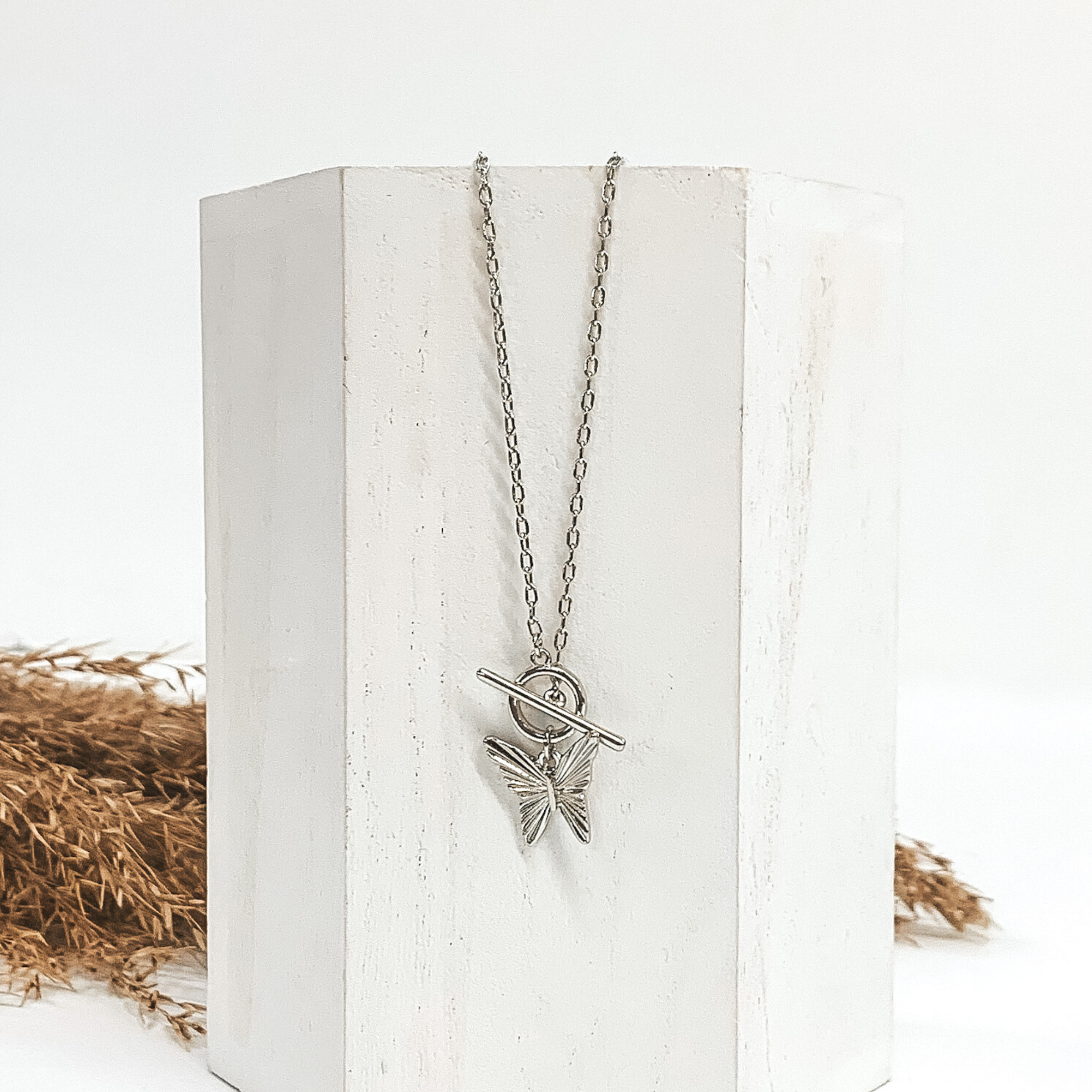 Silver chain necklace with toggle clasp and a hanging silver butterfly pendant. This necklace is laying on a piece of white wood that is in front of a white background with brown floral.