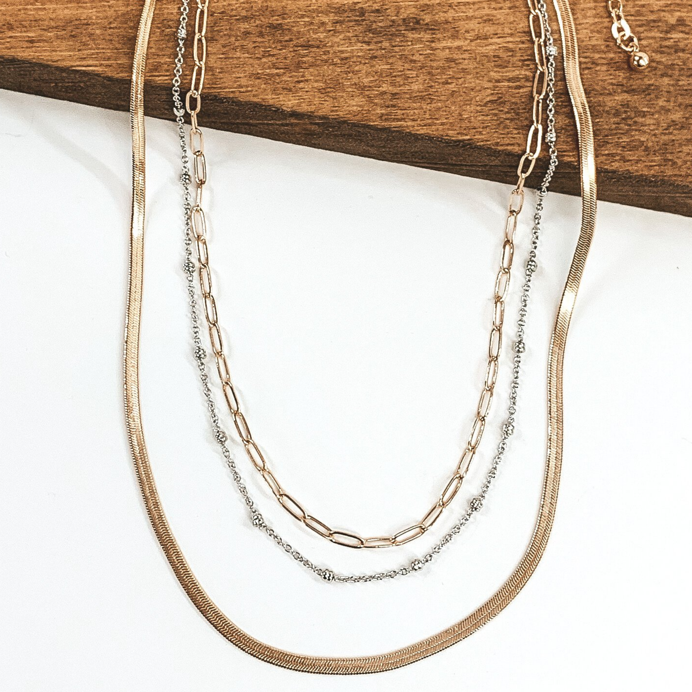 This gold necklace has three different chains. The first one is a classic thin link chain strand in gold, the next one has tiny silver bead spacers on a silver chain, and the last one is a flat herringbone chain. This necklace is pictured on a white background while partially laying on a dark piece of wood.  