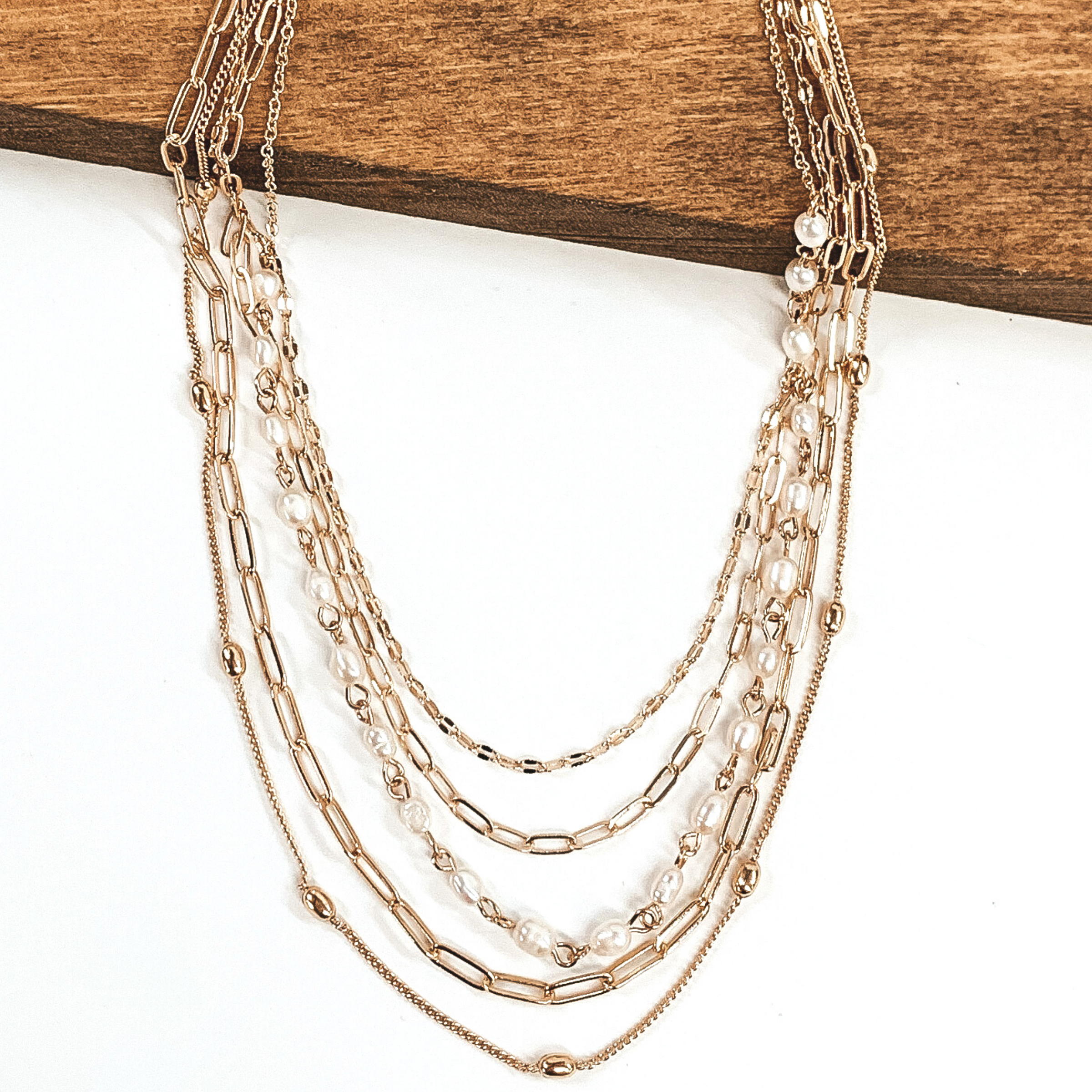 This necklace has 5 different gold chains. One includes gold beads, while another includes white pearls. This necklace is pictured on a white background while partially laying on a dark piece of wood.
