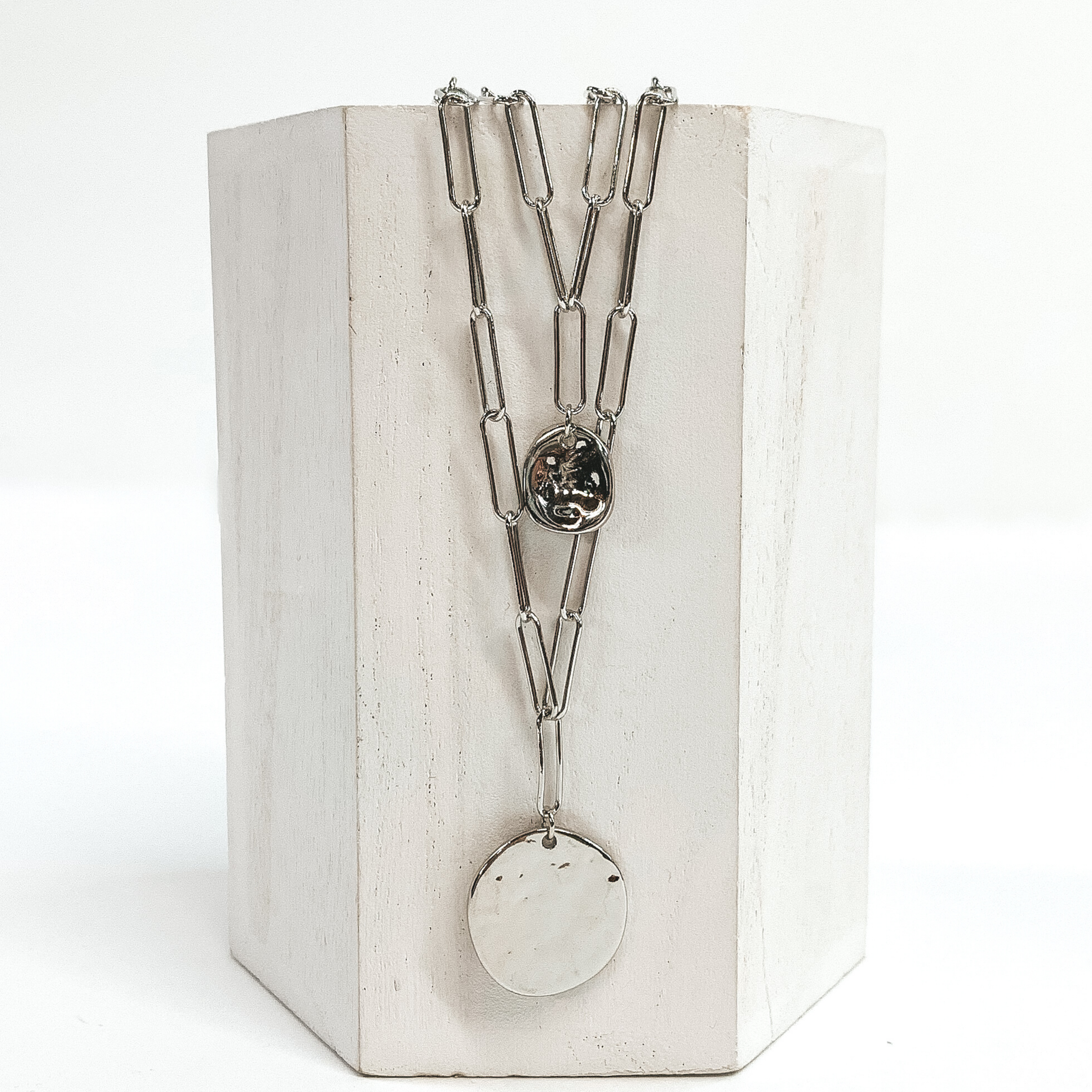 Double layered silver chain necklace with two circle pendants. The pendants have a hammered texture and there is a small one on the shorter strand and a bigger circle on the longer strand. This necklace is pictured on a white block with a white background. 