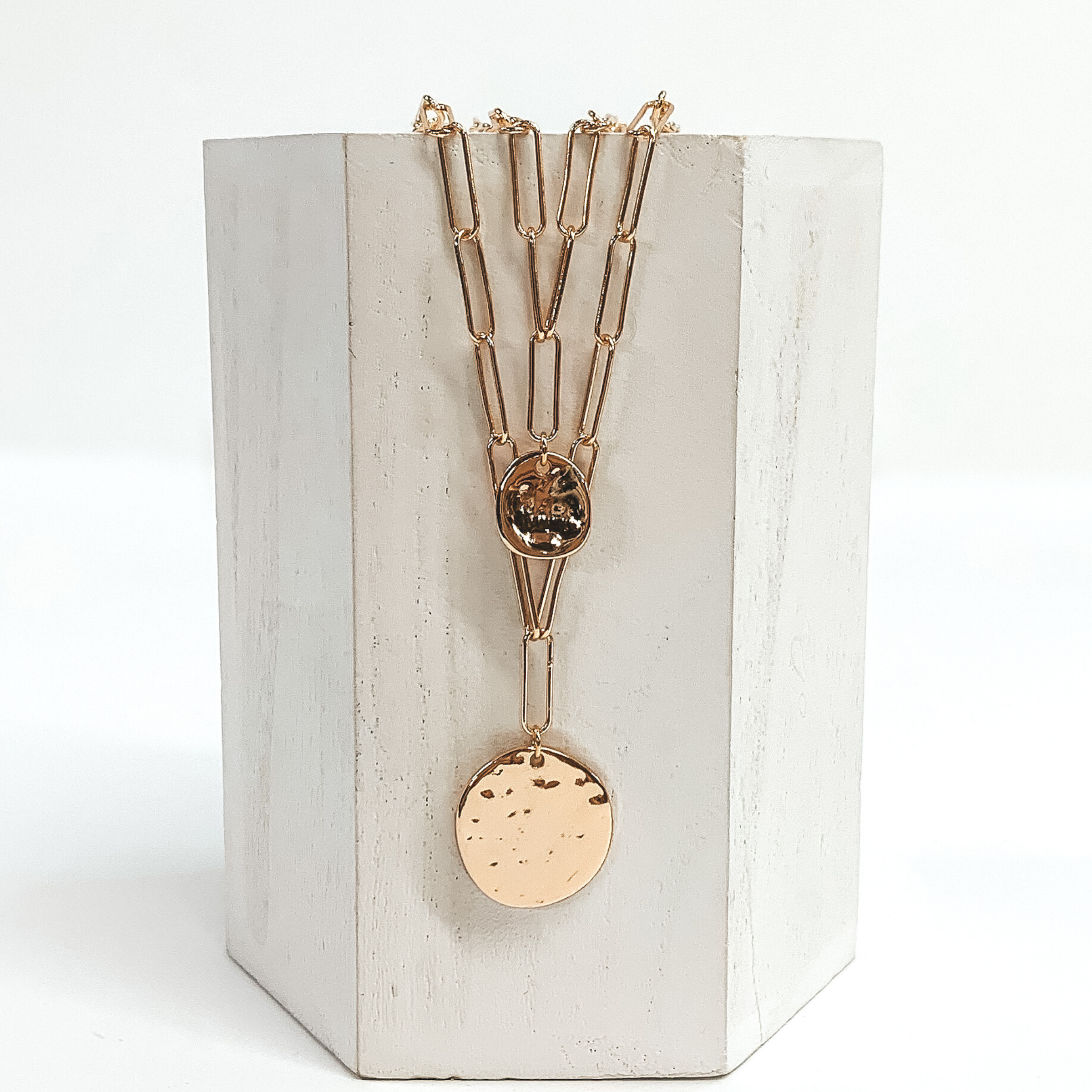 Double layered gold chain necklace with two circle pendants. The pendants have a hammered texture and there is a small one on the shorter strand and a bigger circle on the longer strand. This necklace is pictured on a white block with a white background.