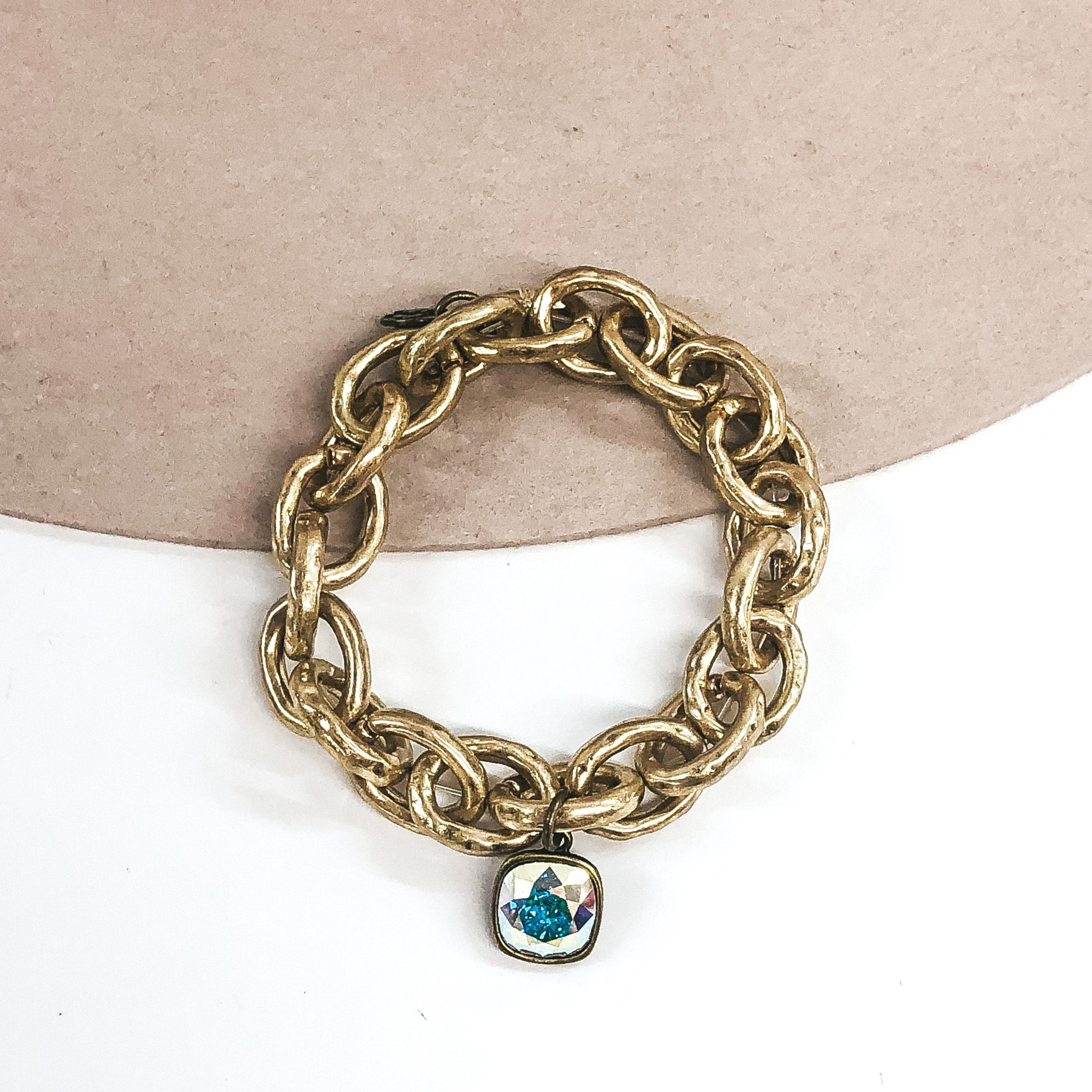 Chunky matte gold chain bracelet with a hanging cushion cut AB crystal that is in a bronze setting. This bracelet is pictured on a beige and white background. 
