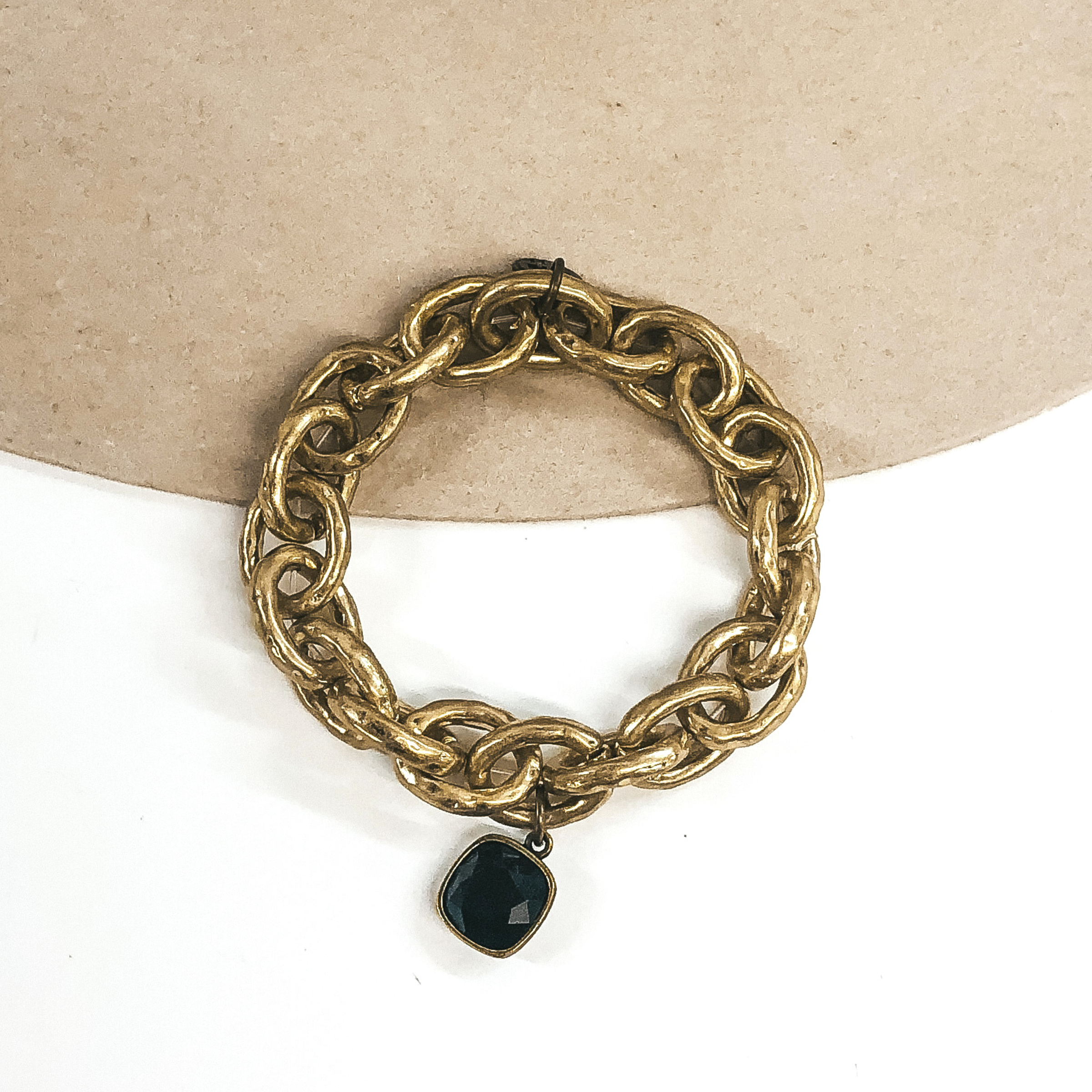 Chunky matte gold chain bracelet with a hanging cushion cut black crystal that is in a bronze setting. This bracelet is pictured on a beige and white background.