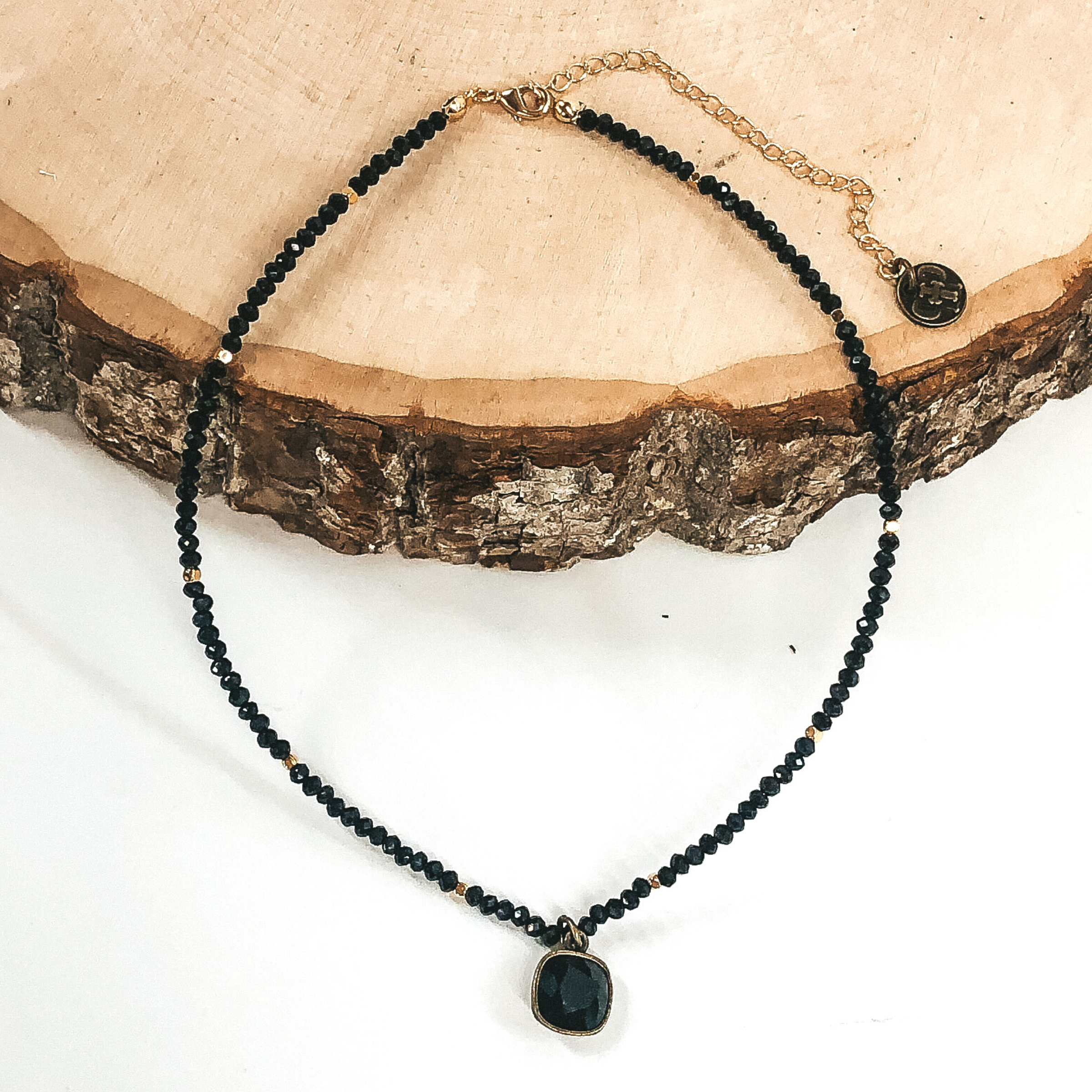This necklace has black crystal beads for the necklace with gold beaded spacers. There is also a single hanging cushion cut black crystal in a bronze setting. This necklace is pictured laying partially on a piece of wood on a white background.