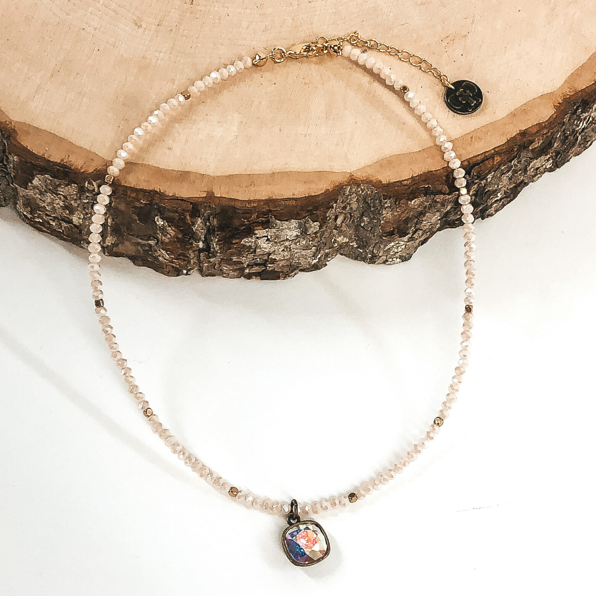 This necklace has blush crystal beads for the necklace with gold beaded spacers. There is also a single hanging cushion cut AB crystal in a bronze setting. This necklace is pictured laying partially on a piece of wood on a white background. 