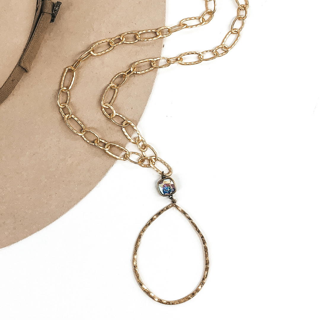 Gold chain link necklace with a open hammered teardrop pendant with a ab cushion cut crystal connector. This necklace is laying partially on a beige hat that is on a white background. 