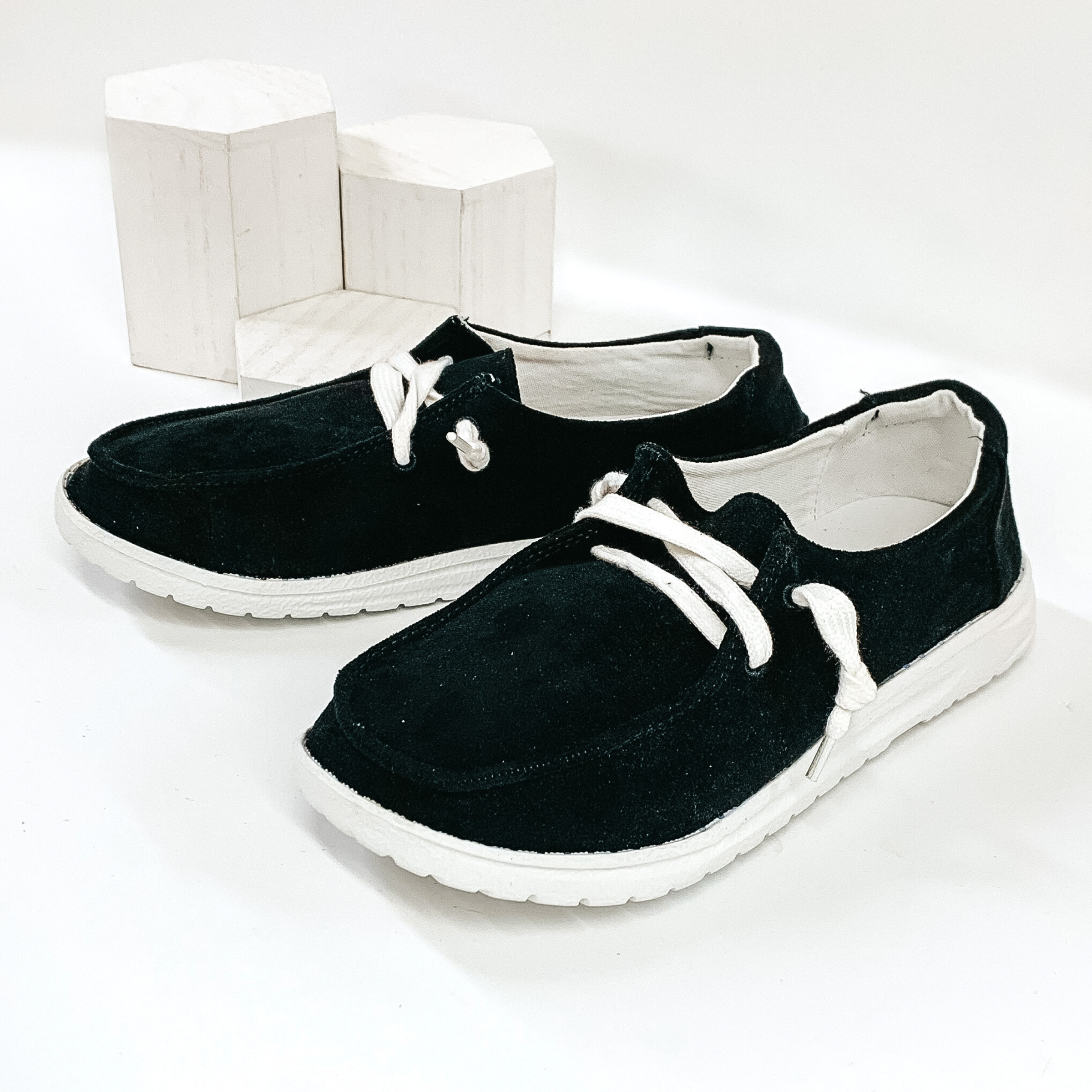 Black slip on suede loafers with a white colored shoe lace and white sole. Pictured on white background.
