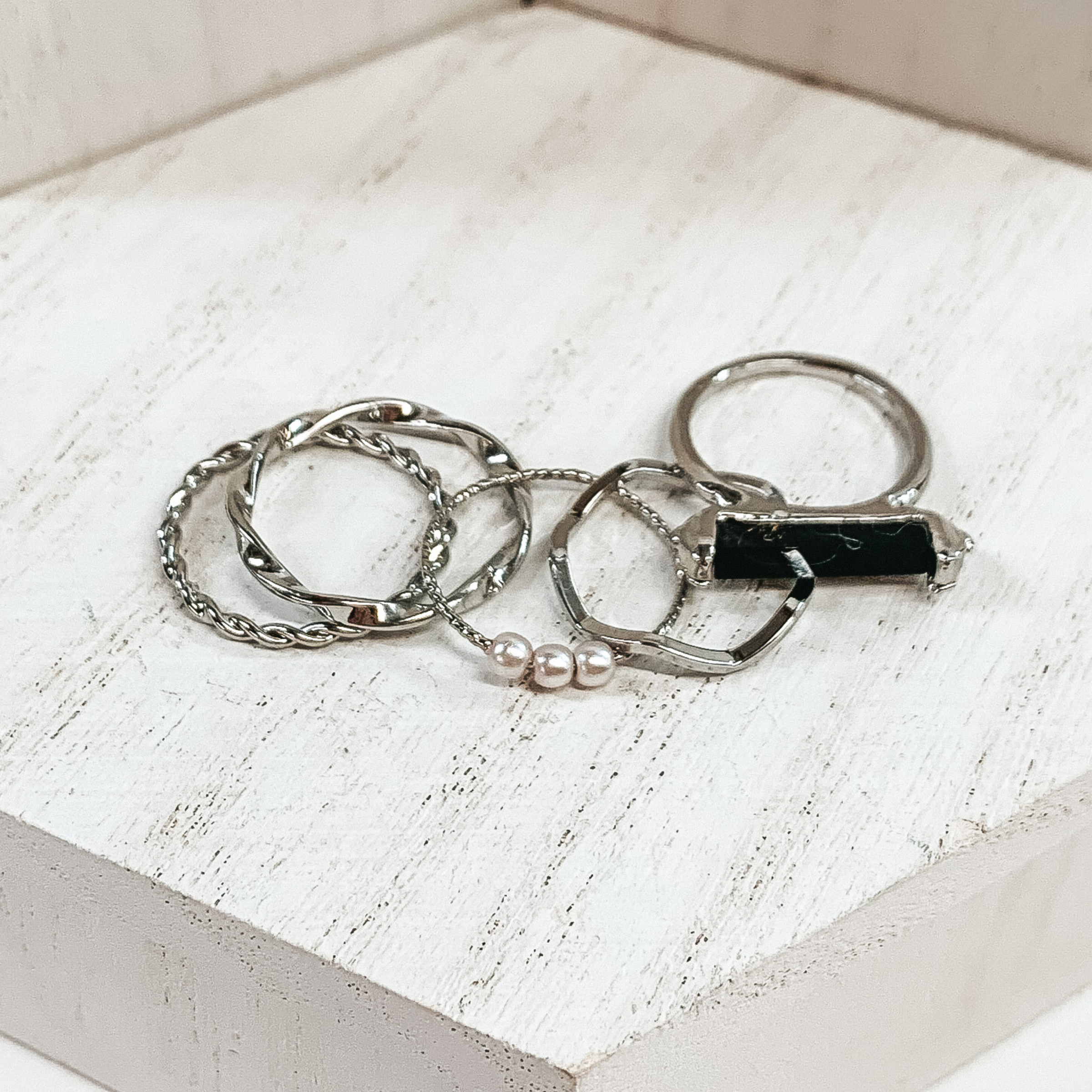 This is a set of five silver rings. You have two twisted rings, one wavy ring, on ring that has 3 pearls, and the last ring has a black bar stone. These rings are pictured on a white background. 