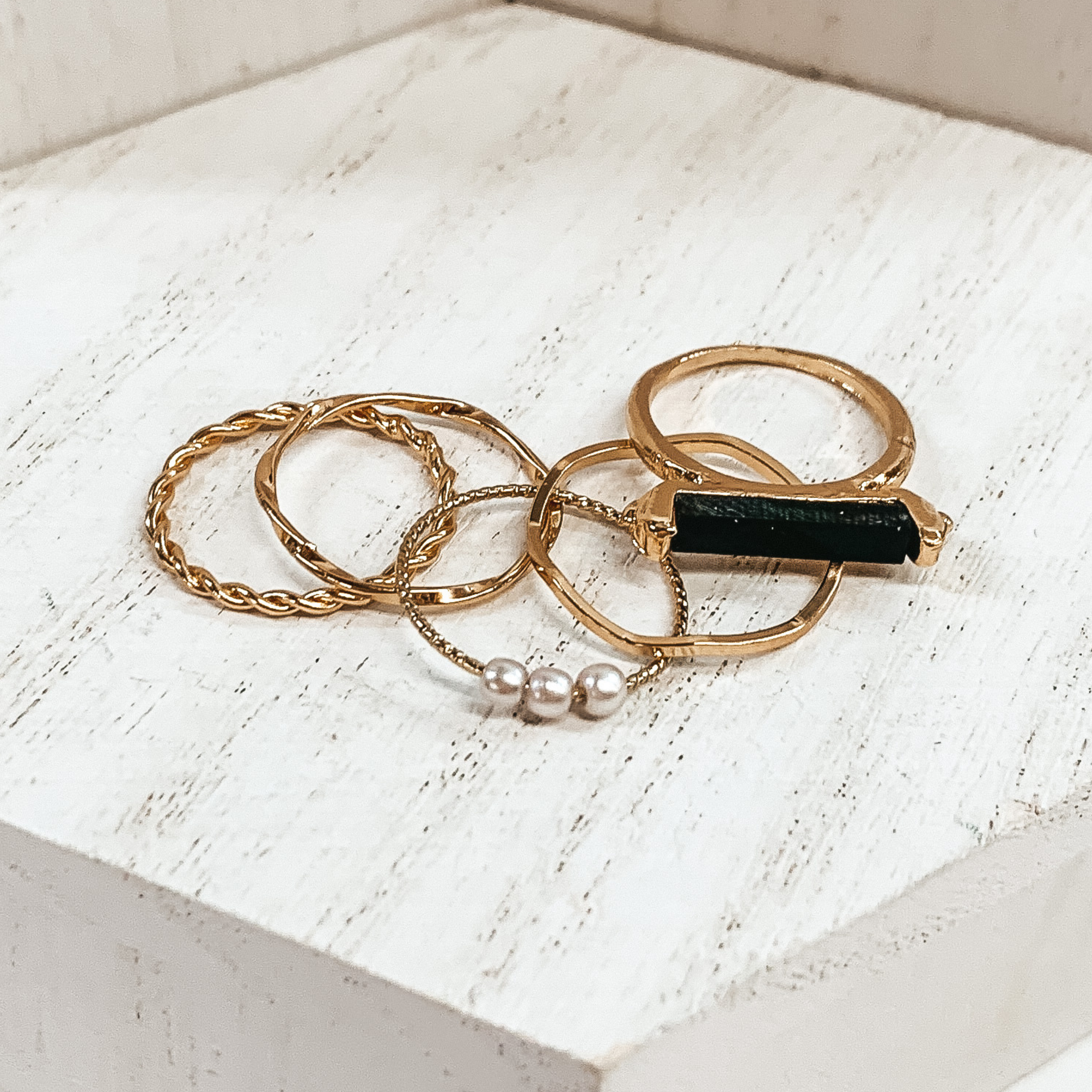 This is a set of five gold rings. You have two twisted rings, one wavy ring, on ring that has 3 pearls, and the last ring has a black bar stone. These rings are pictured on a white background.