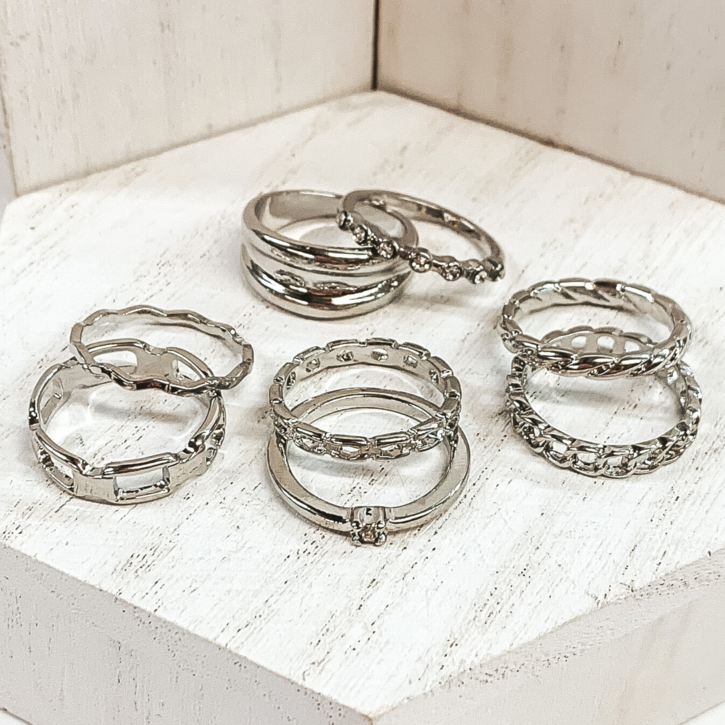 This is a set of eight silver rings. Two of the rings include clear crystals, three rings are different types of chains, one twisted ring, one wavy rings, and one thicker banded ring. This set is pictured on a white background.