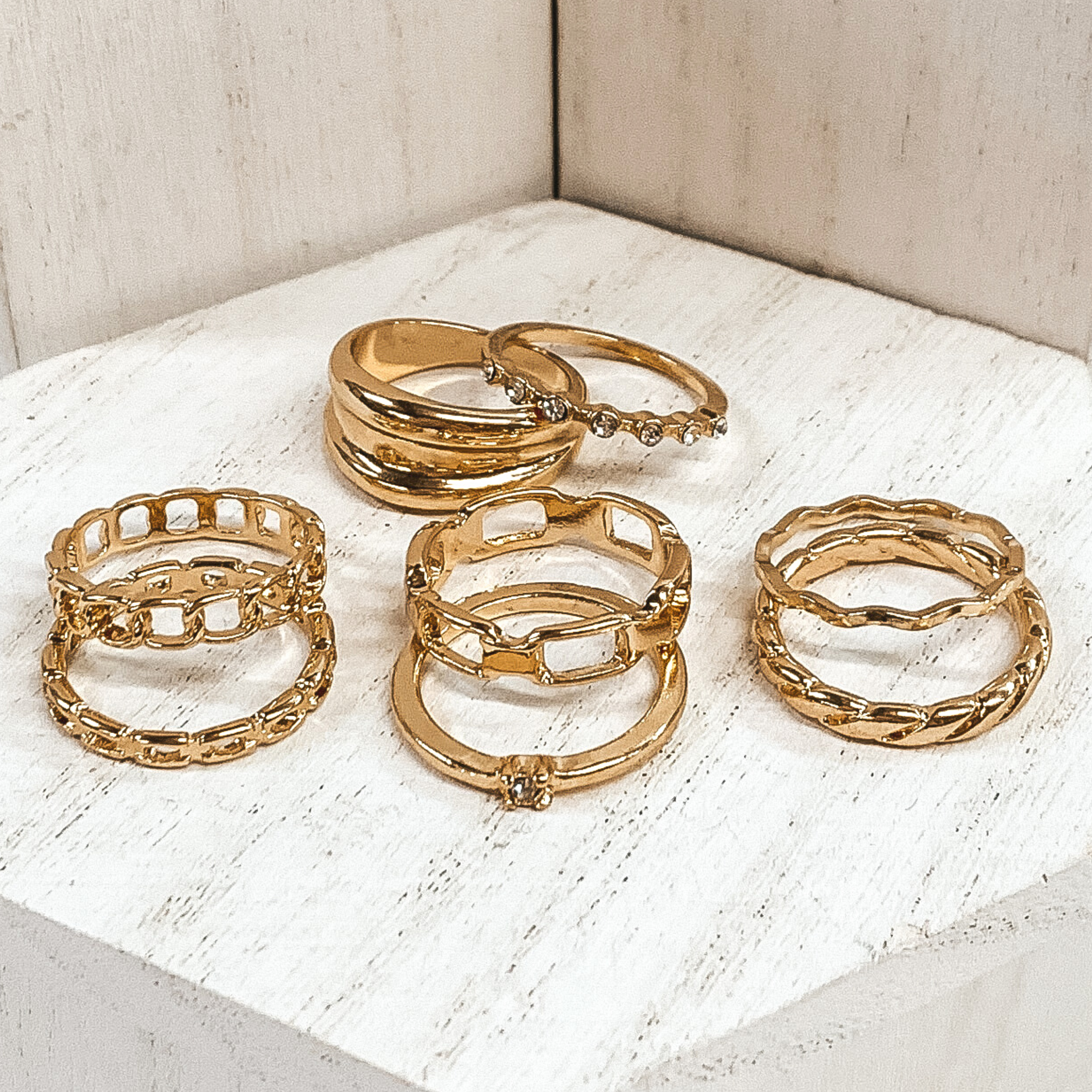 This is a set of eight gold rings. Two of the rings include clear crystals, three rings are different types of chains, one twisted ring, one wavy rings, and one thicker banded ring. This set is pictured on a white background. 