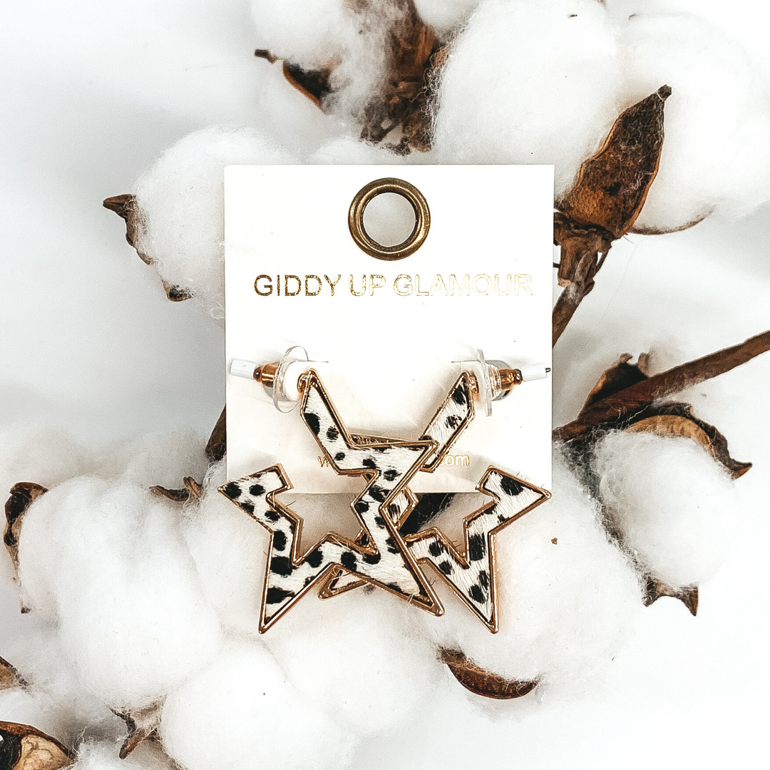 Gold outline star hoop earrings with a white hide inlay with black dots. These earrings are pictured laying on white cotton on a white background.