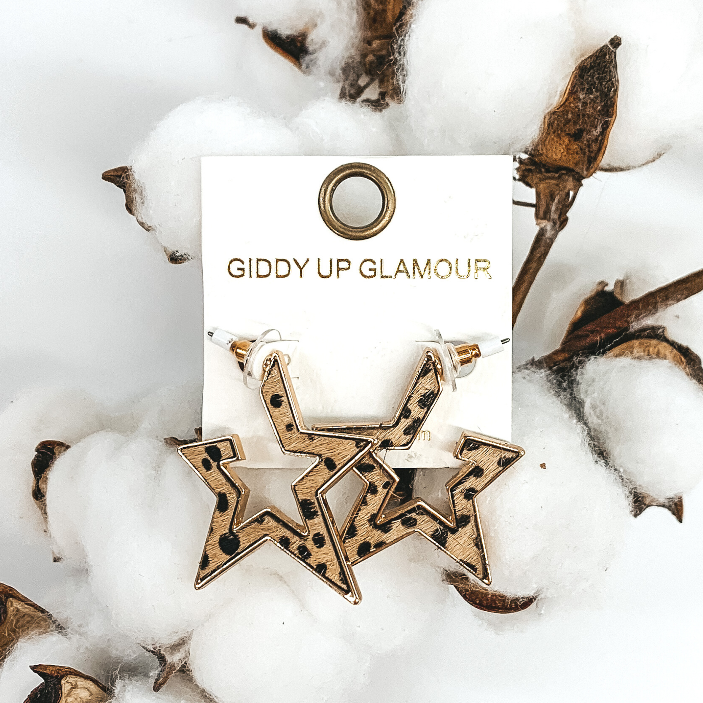 Gold outline star hoop earrings with a brown hide inlay with black dots. These earrings are pictured laying on white cotton on a white background.
