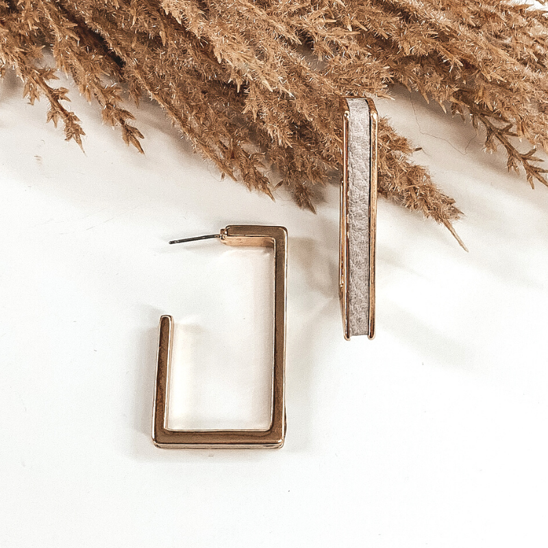 Gold outline rectangle hoop earrings with a grey leather material inlay. These earrings are pictured laying on white background with brown floral at the top of the picture. 