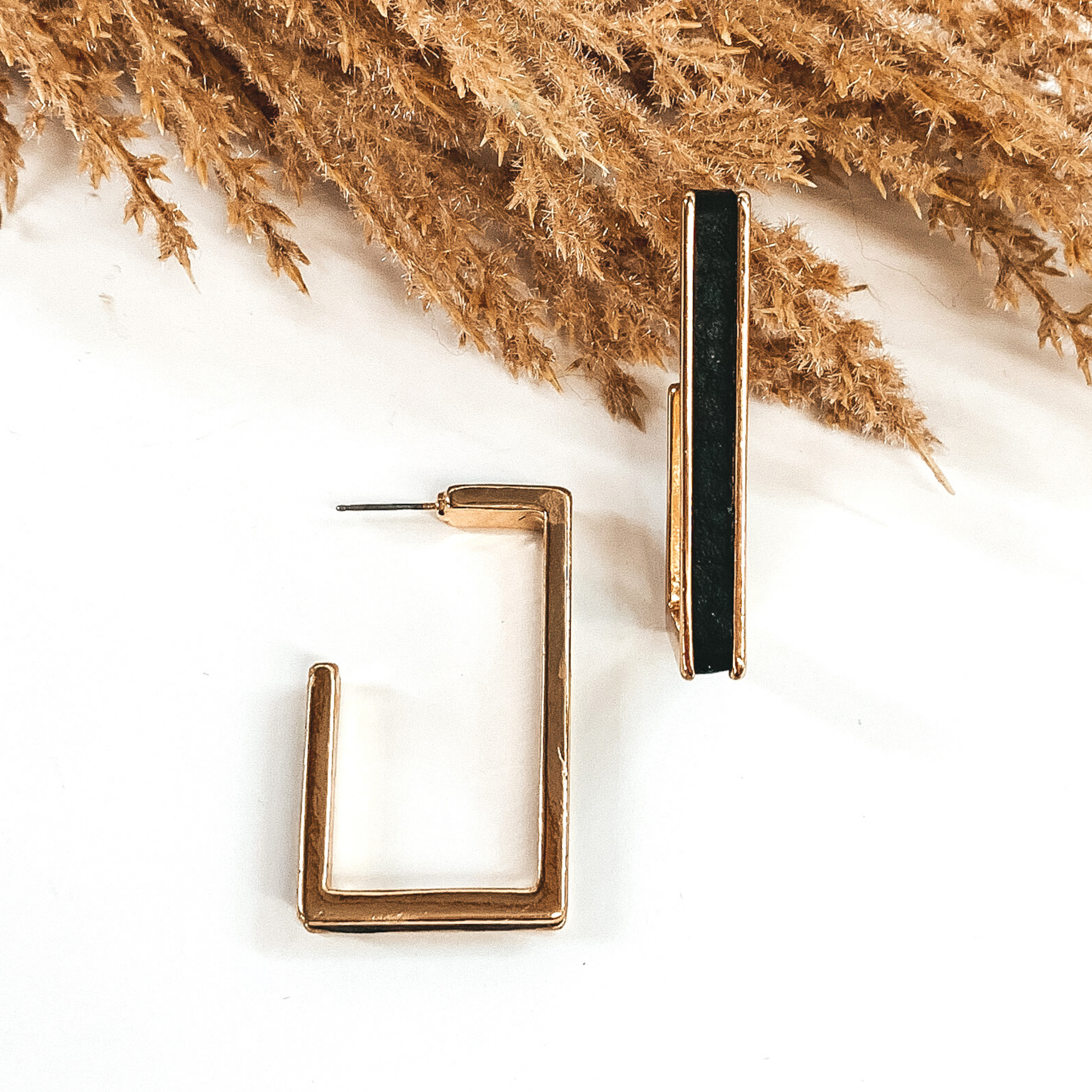 Gold outline rectangle hoop earrings with a black leather material inlay. These earrings are pictured laying on white background with brown floral at the top of the picture. 