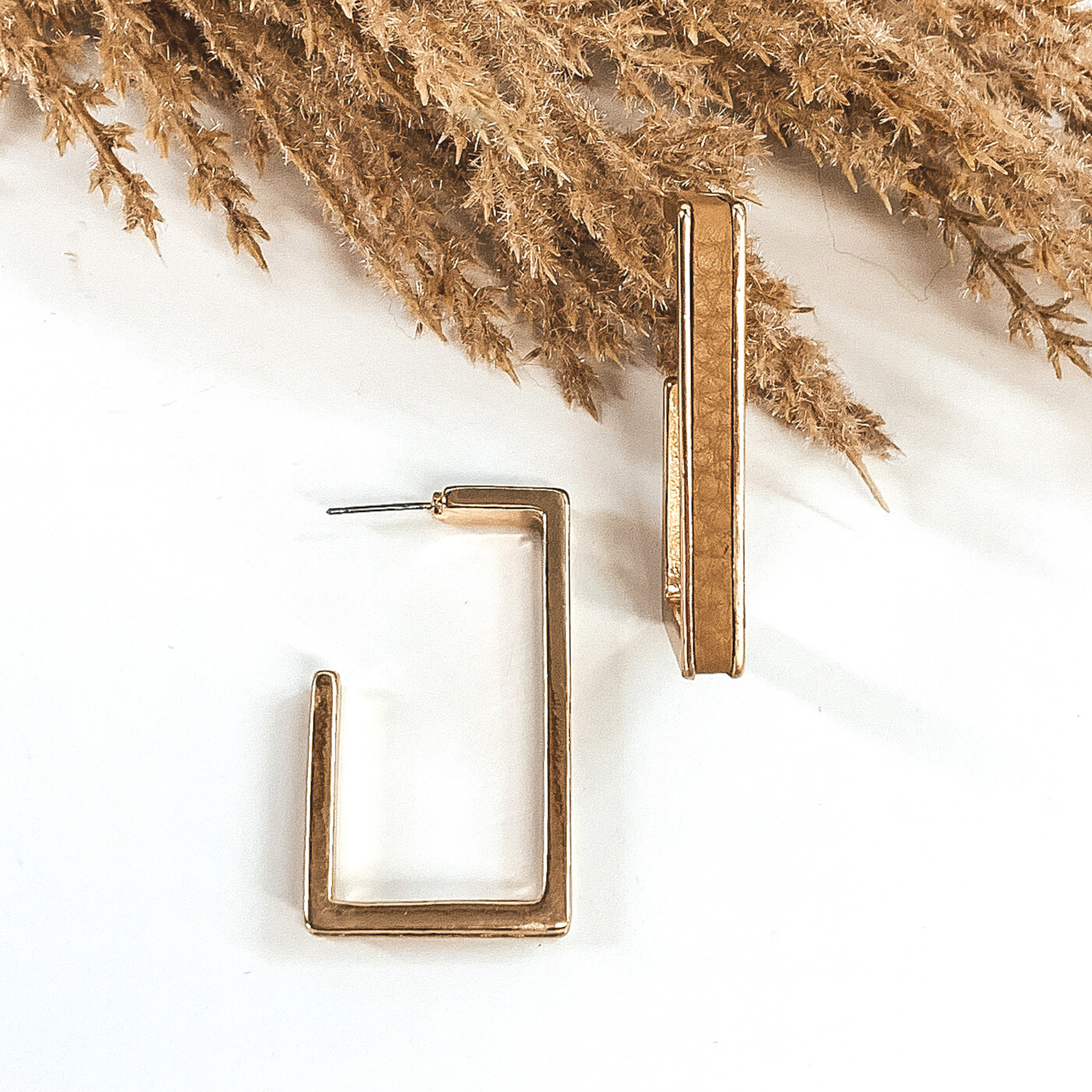 Gold outline rectangle hoop earrings with a tan leather material inlay. These earrings are pictured laying on white background with brown floral at the top of the picture. 