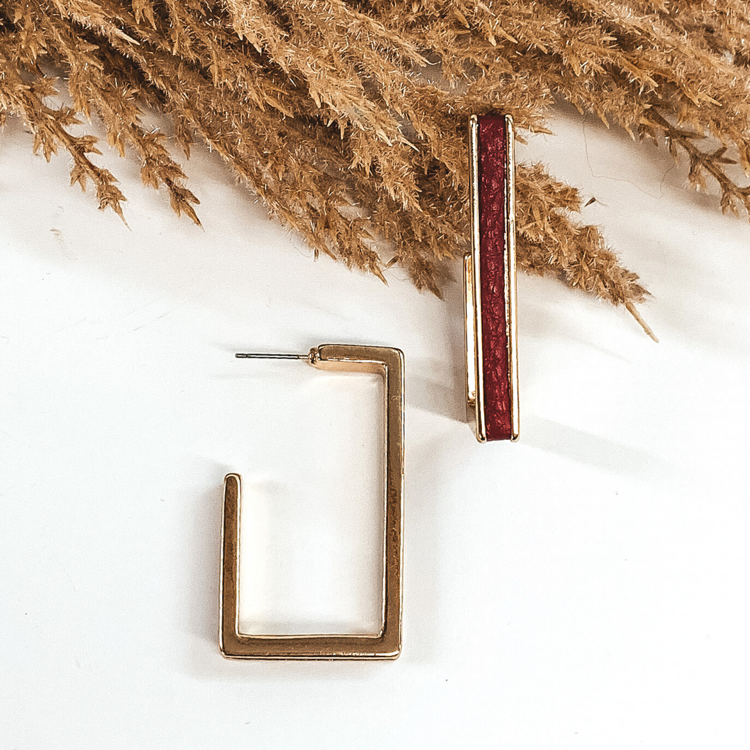 Gold outline rectangle hoop earrings with a red leather material inlay. These earrings are pictured laying on white background with brown floral at the top of the picture. 