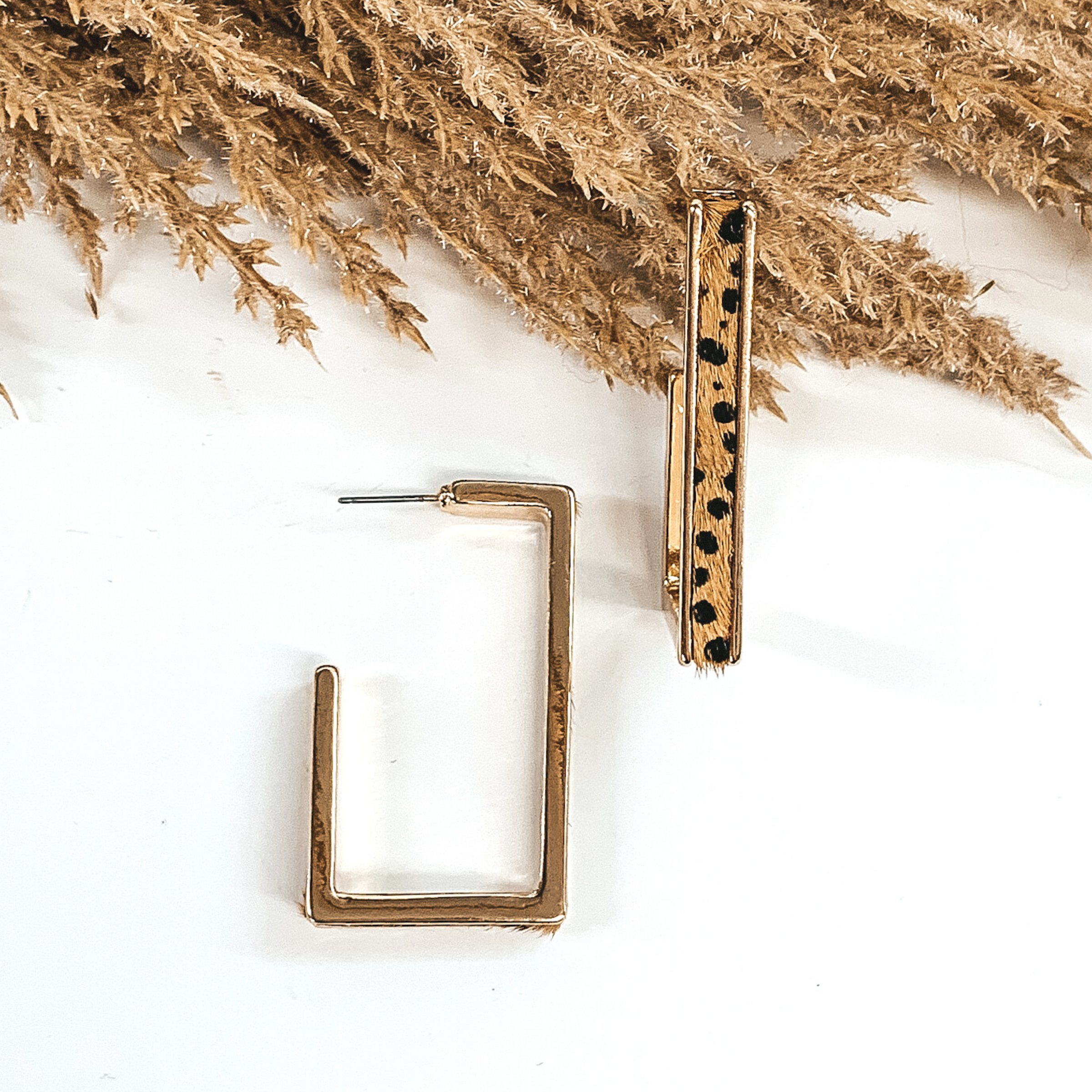 Gold outline rectangle hoop earrings with a brown dotted cow hide leather inlay. These earrings are pictured laying on white background with brown floral at the top of the picture. 