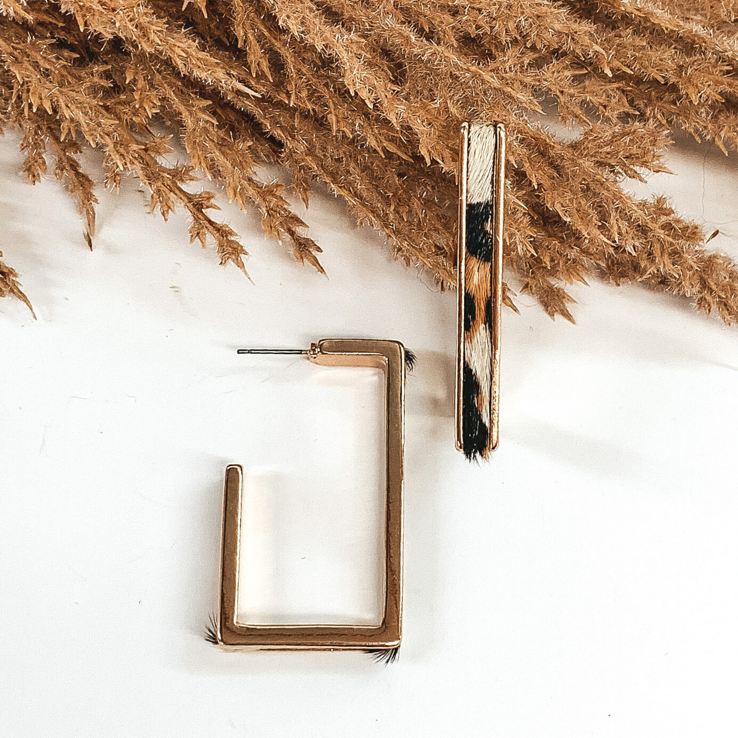 Gold outline rectangle hoop earrings with a white leopard cow hide leather inlay. These earrings are pictured laying on white background with brown floral at the top of the picture. 