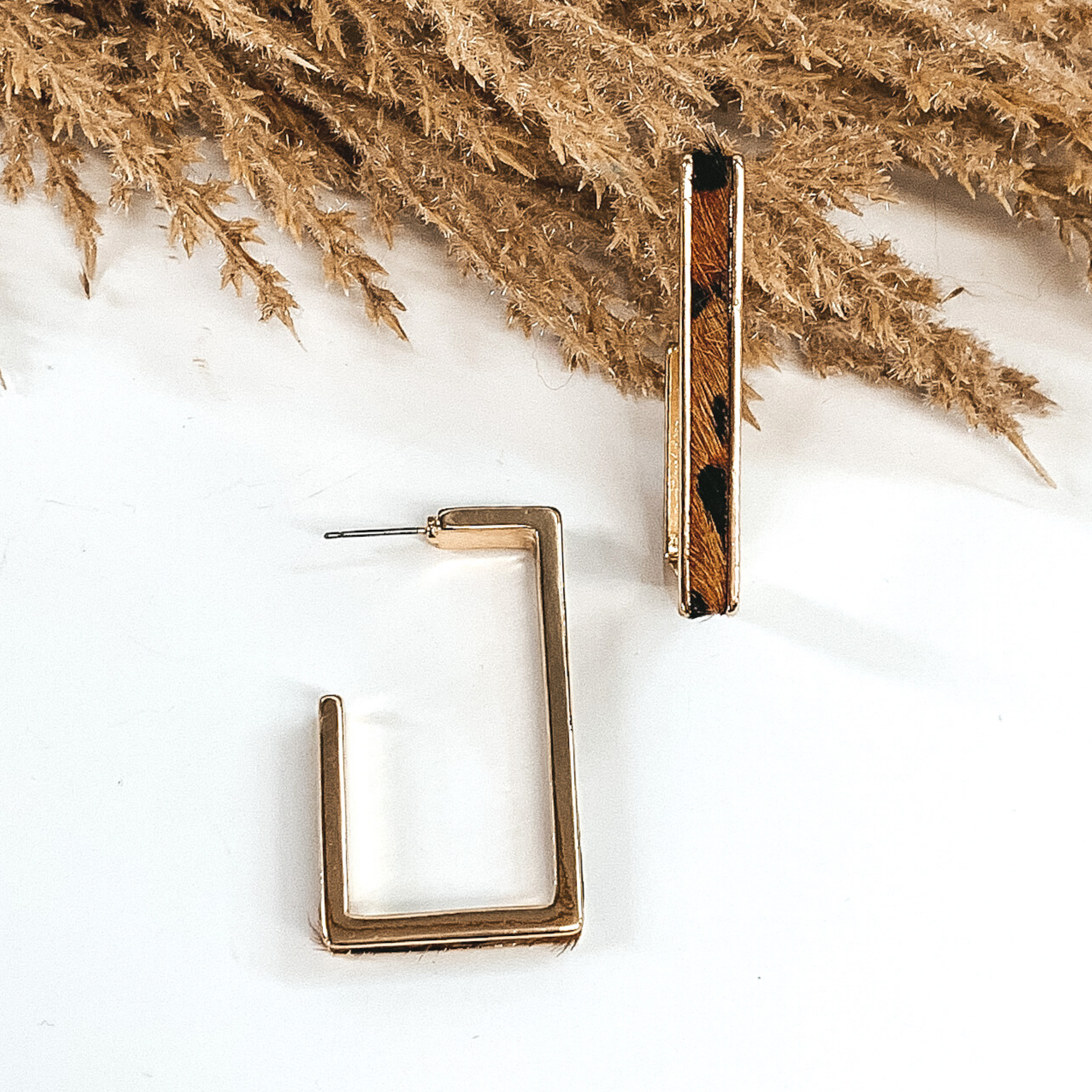 Gold outline rectangle hoop earrings with a brown leopard cow hide leather inlay. These earrings are pictured laying on white background with brown floral at the top of the picture.