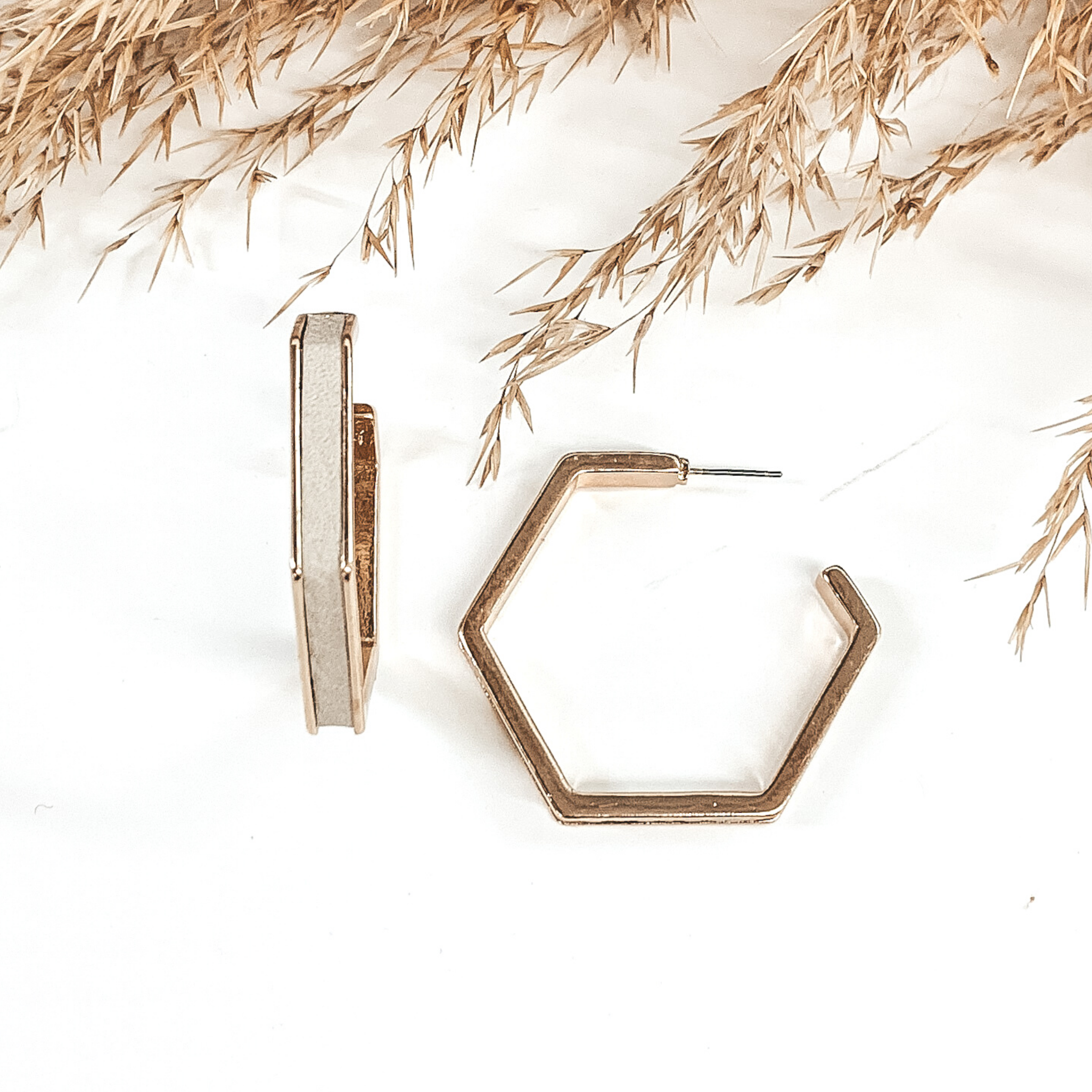 Gold outline hexagon hoop earrings with a grey leather material inlay. These earrings are pictured laying on white background with brown floral at the top of the picture. 