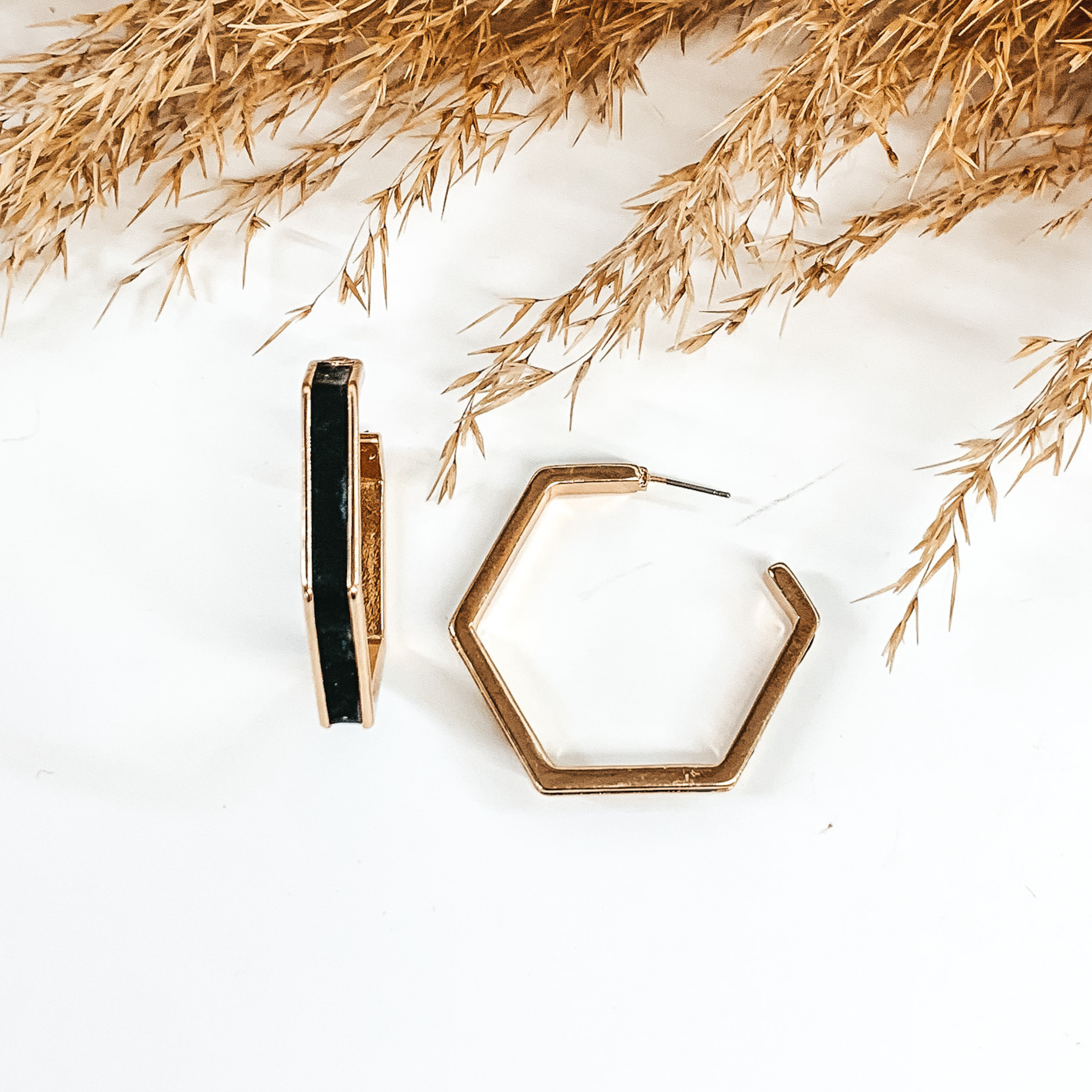 Gold outline hexagon hoop earrings with a black leather material inlay. These earrings are pictured laying on white background with brown floral at the top of the picture. 