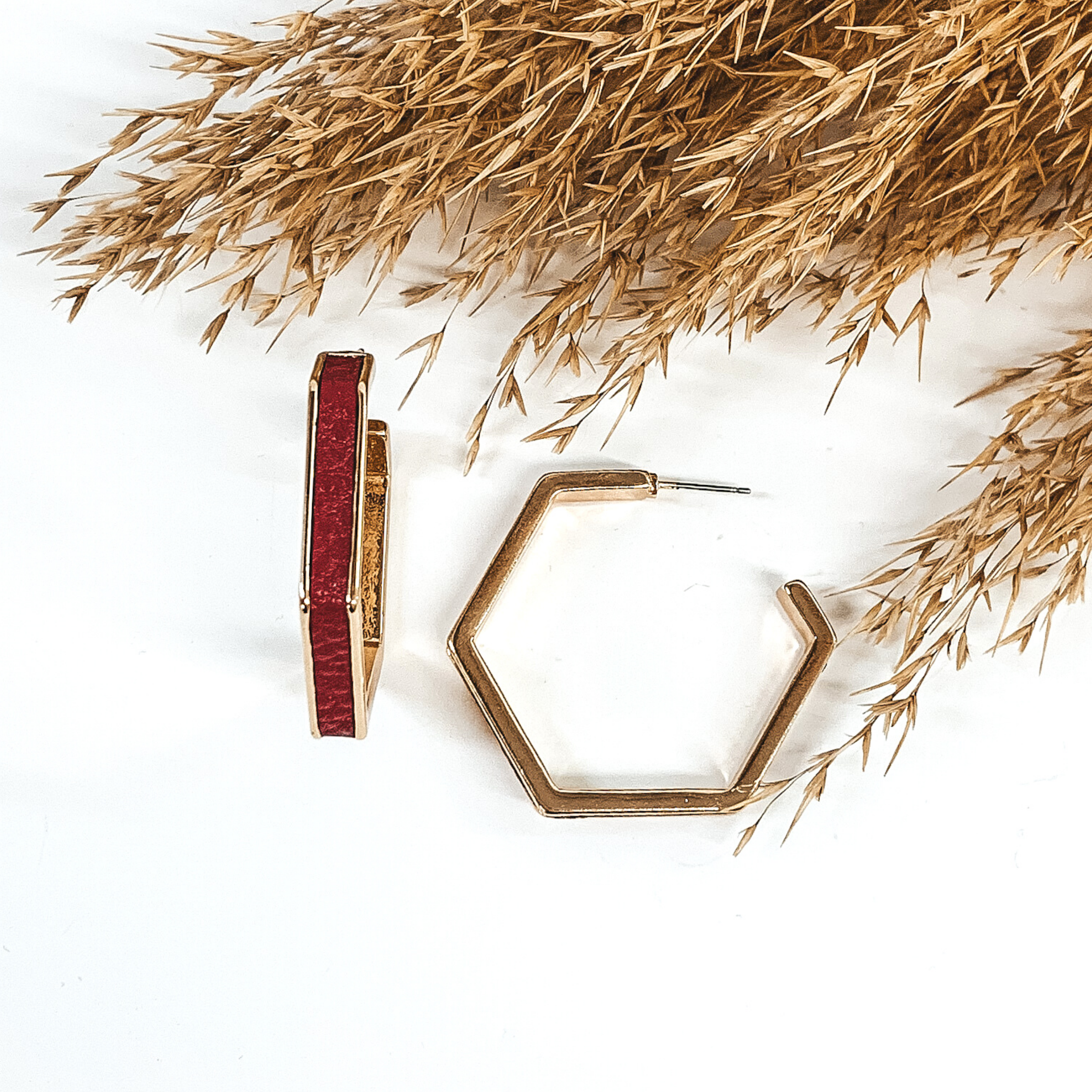 Gold outline hexagon hoop earrings with a red leather material inlay. These earrings are pictured laying on white background with brown floral at the top of the picture. 