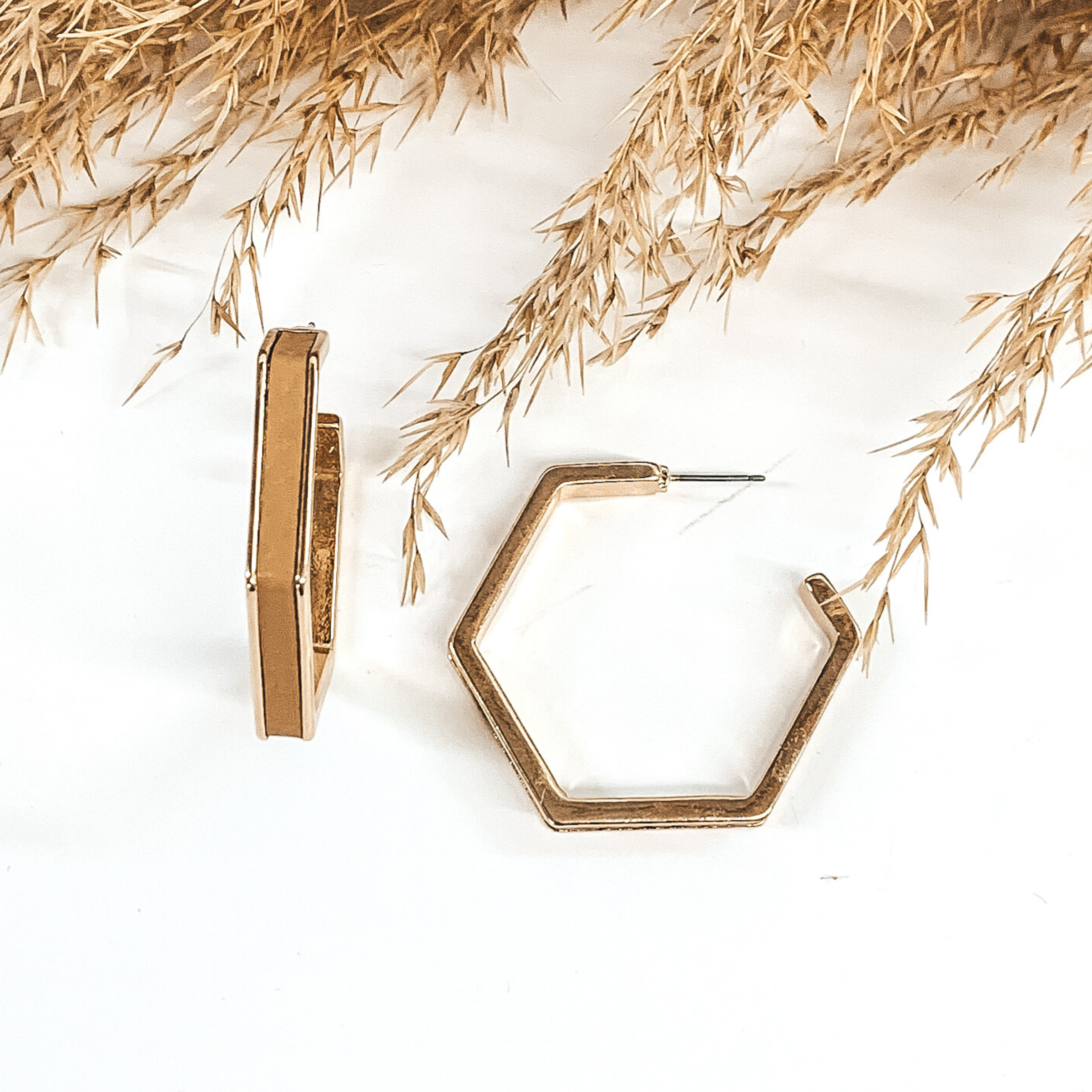 Gold outline hexagon hoop earrings with a tan leather material inlay. These earrings are pictured laying on white background with brown floral at the top of the picture. 