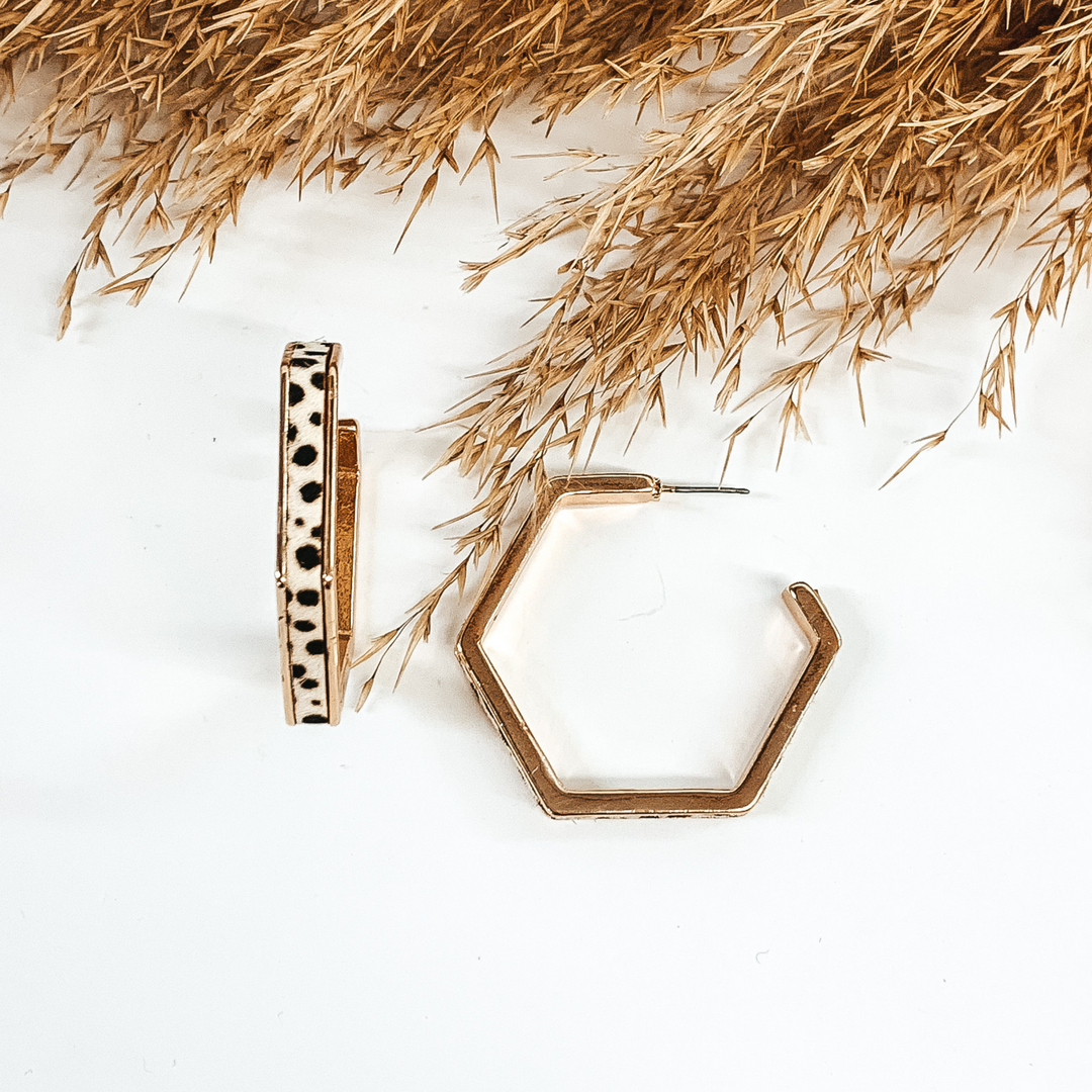 Gold outline hexagon hoop earrings with white dotted cow hide inlay. These earrings are pictured laying on white background with brown floral at the top of the picture. 