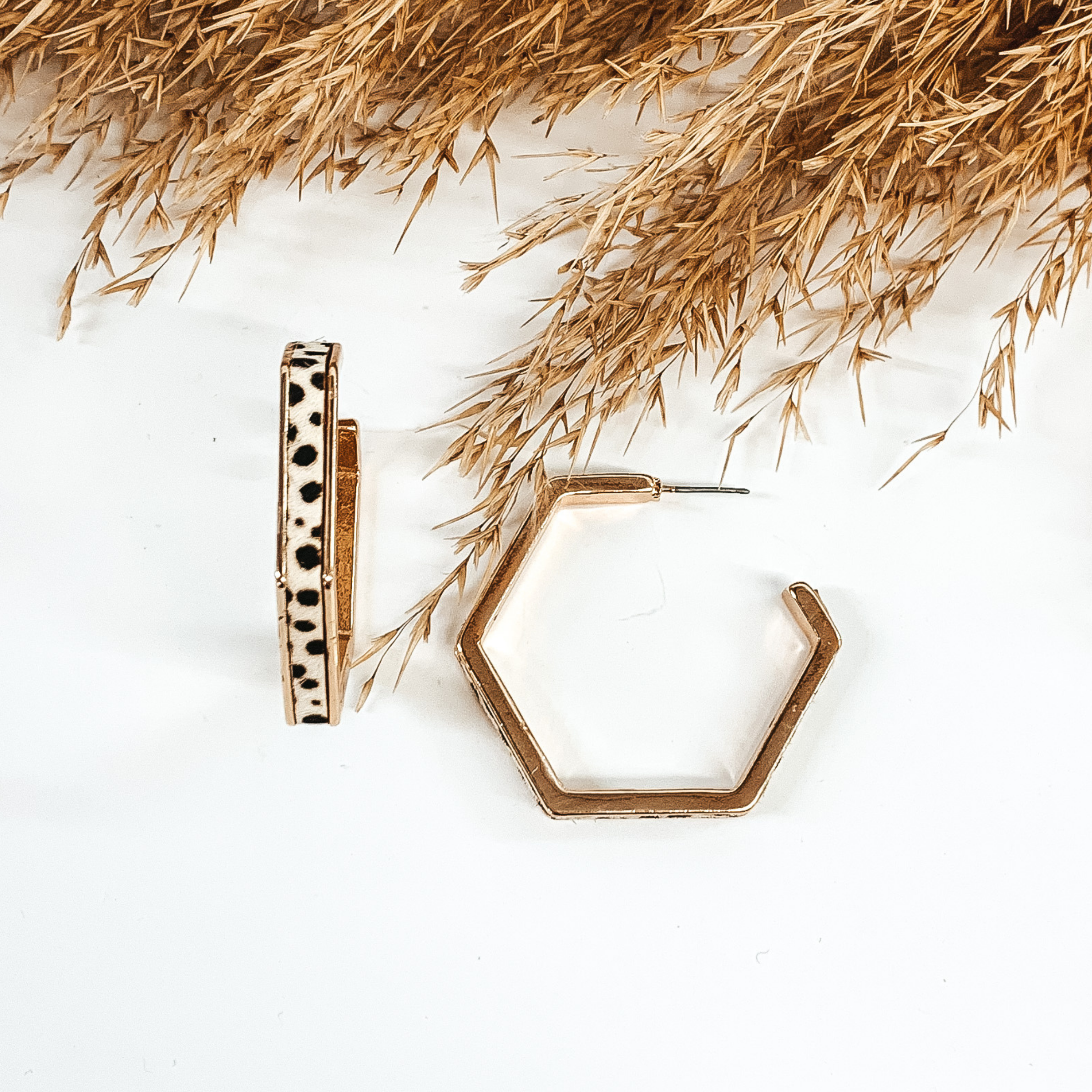 Gold outline hexagon hoop earrings with white dotted cow hide inlay. These earrings are pictured laying on white background with brown floral at the top of the picture. 