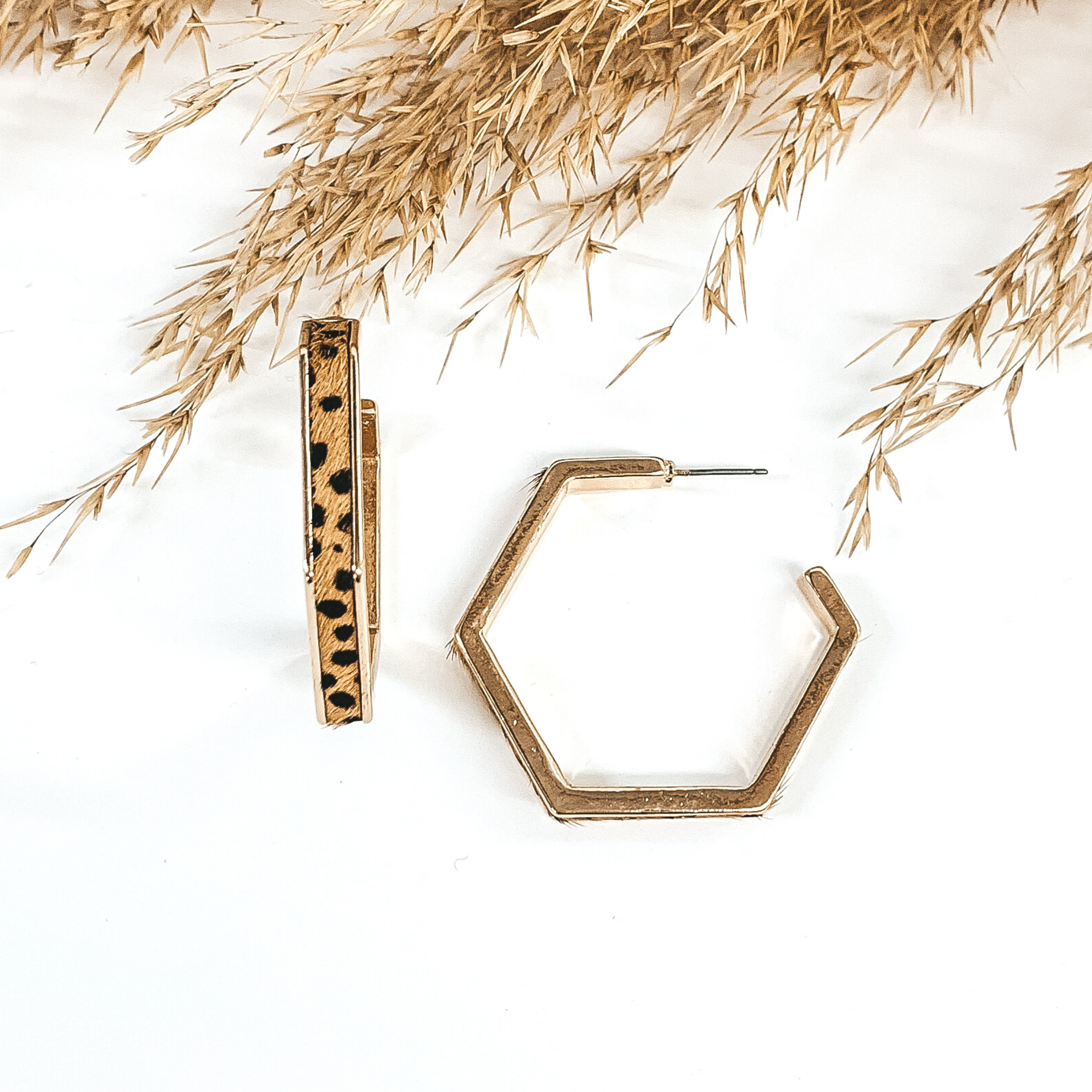 Gold outline hexagon hoop earrings with brown dotted cow hide inlay. These earrings are pictured laying on white background with brown floral at the top of the picture. 