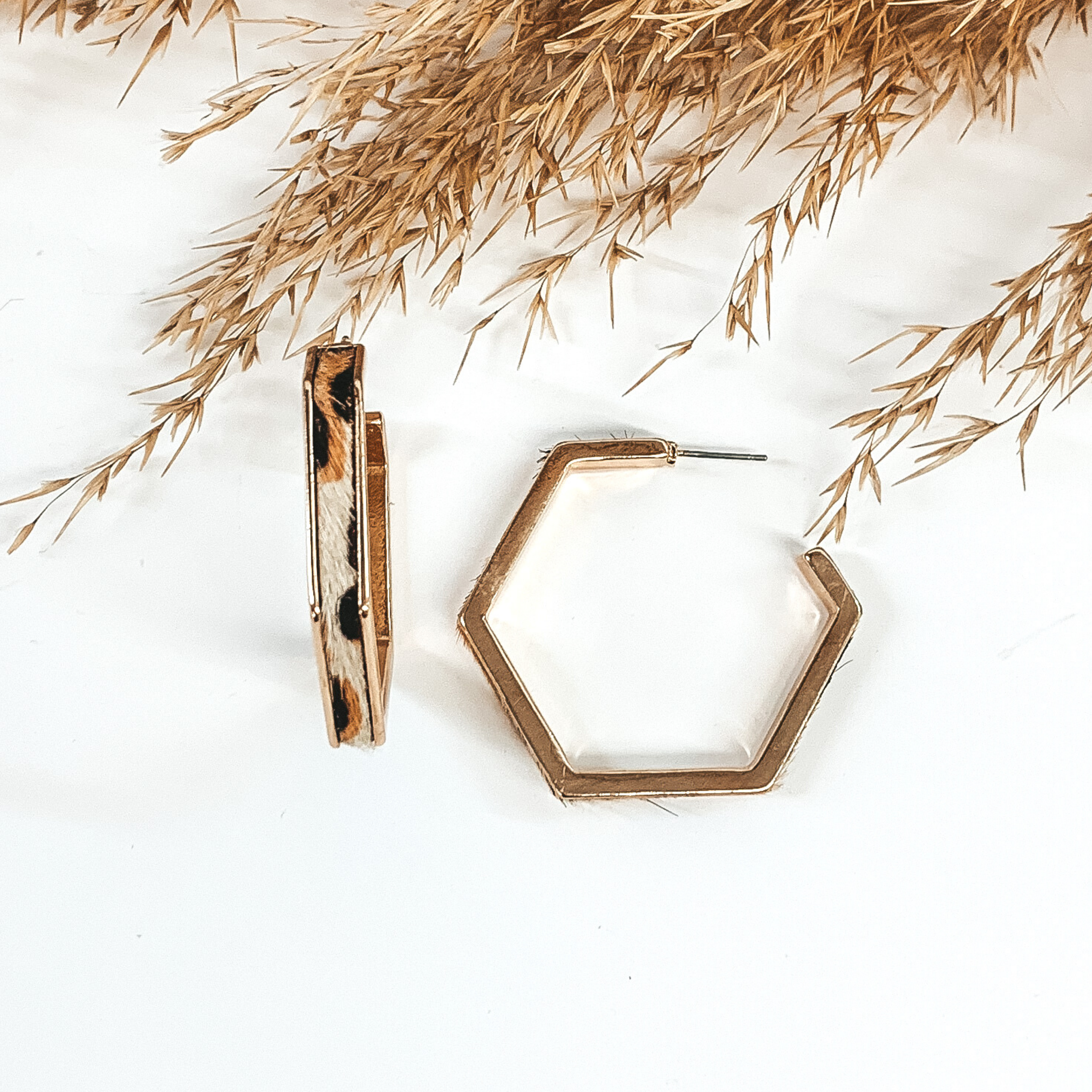 Gold outline hexagon hoop earrings with white leopard cow hide inlay. These earrings are pictured laying on white background with brown floral at the top of the picture. 