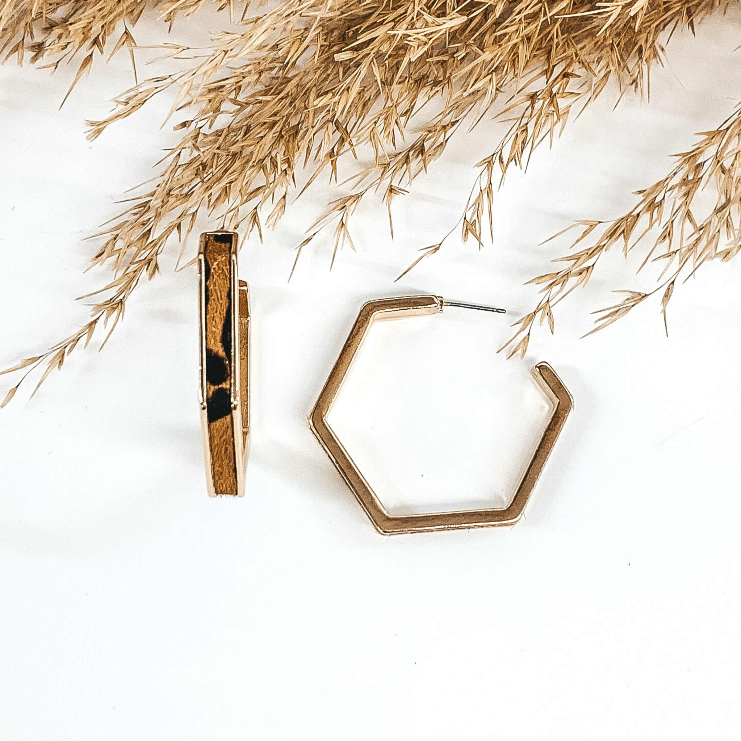 Gold outline hexagon hoop earrings with brown leopard cow hide inlay. These earrings are pictured laying on white background with brown floral at the top of the picture. 