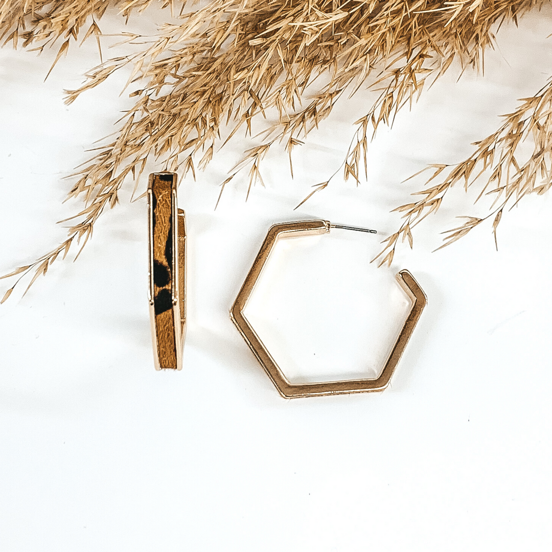 Gold outline hexagon hoop earrings with brown leopard cow hide inlay. These earrings are pictured laying on white background with brown floral at the top of the picture. 