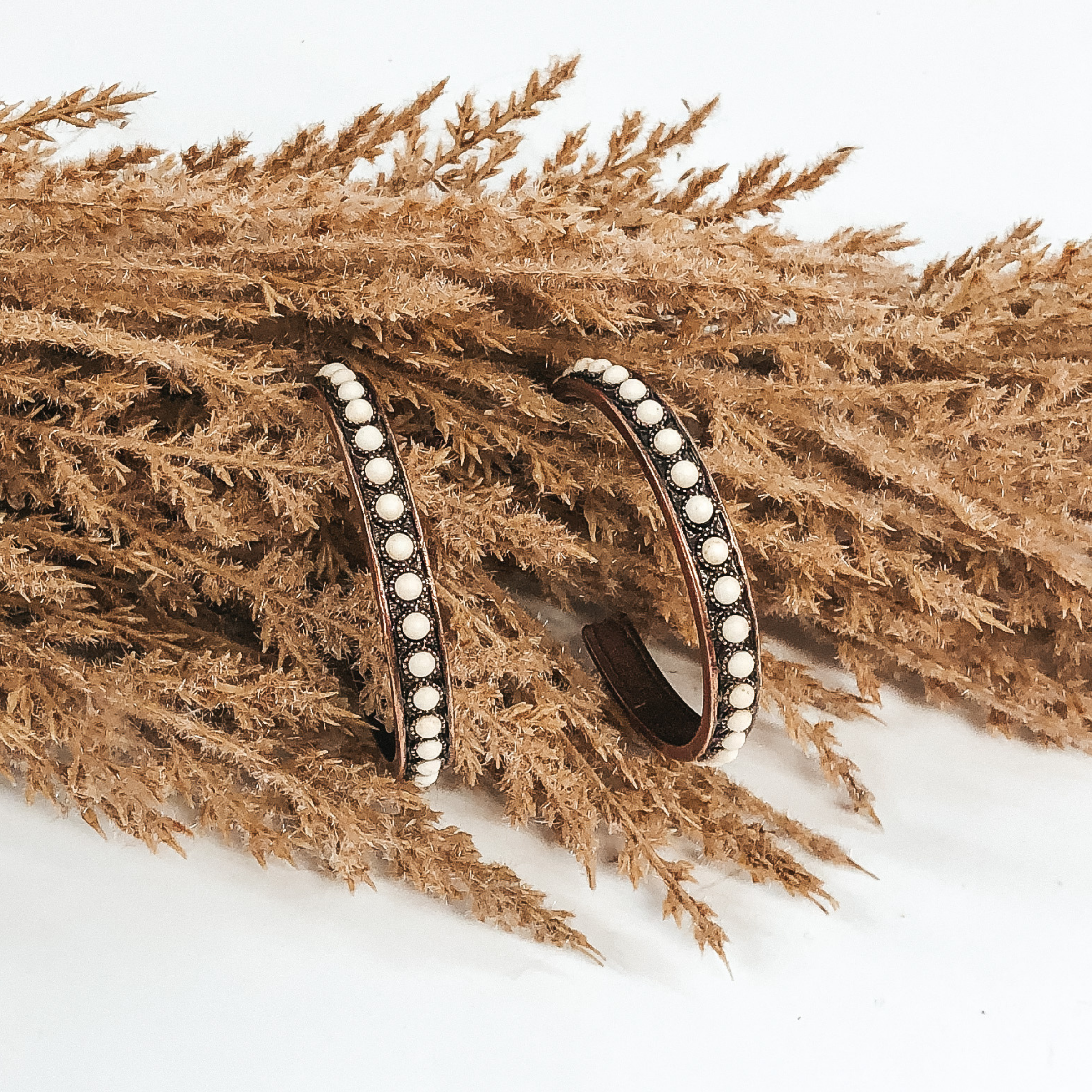 Copper colored hoops with white circular stones covering the hoops in a line. These hoops are pictured hanging on brown floral that is on a white background. 