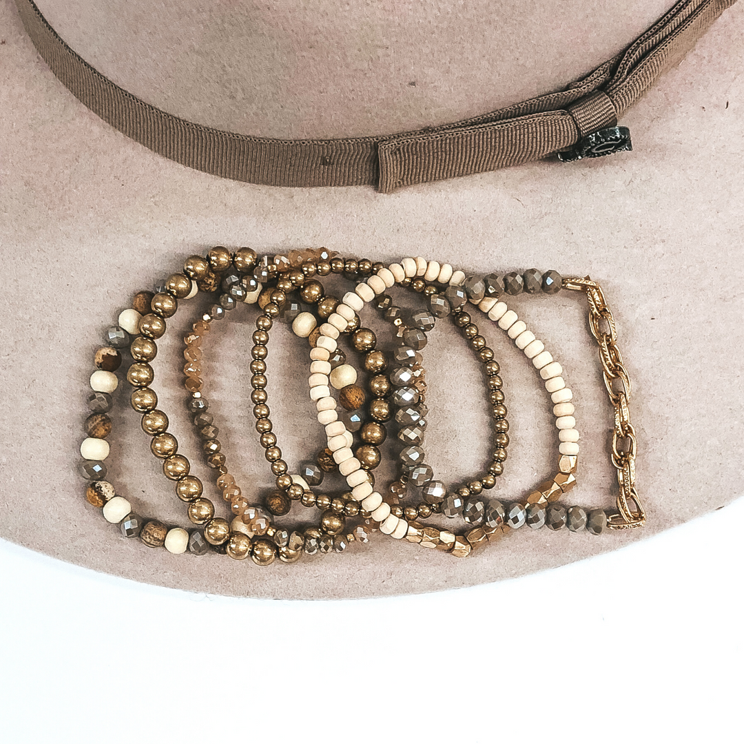 This bracelet set has 6 different beaded bracelets. One has different hues of brown beads, you have two gold beads (one is small and the other is bigger), one bracelet has two different hues of brown crystal beads that has gold spacers, one has brown crystal beads with a gold chain on one half of it, and the last bracelet has ivory and gold beads. This bracelet set is pictured on a beige hat on a white background.