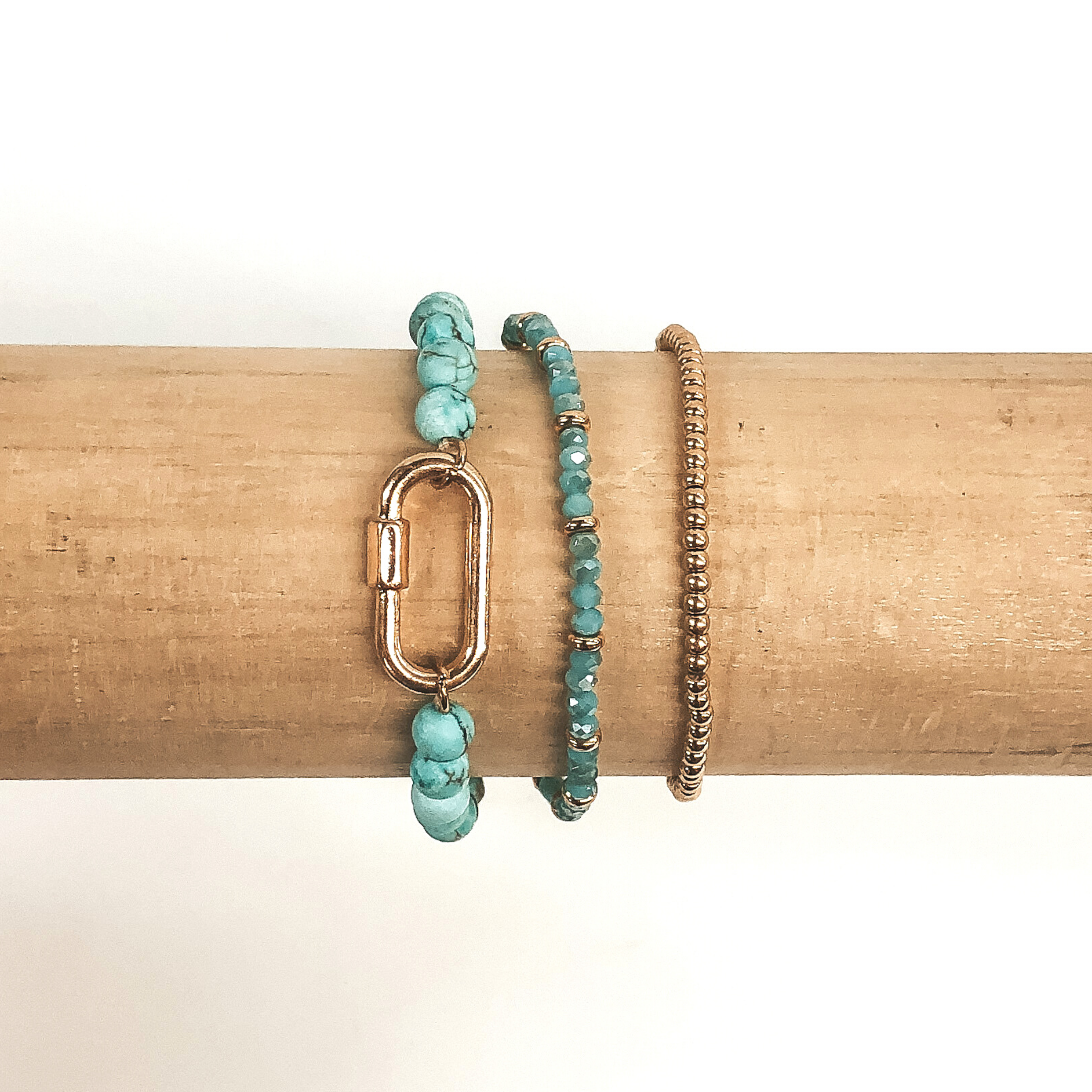 One bracelet is all gold beads, one bracelet has dark teal beads and gold spacer beads, and the last bracelet has teal marble beads with an oval pendant. These bracelets are pictured on a wood colored bracelet holder on a white background. 