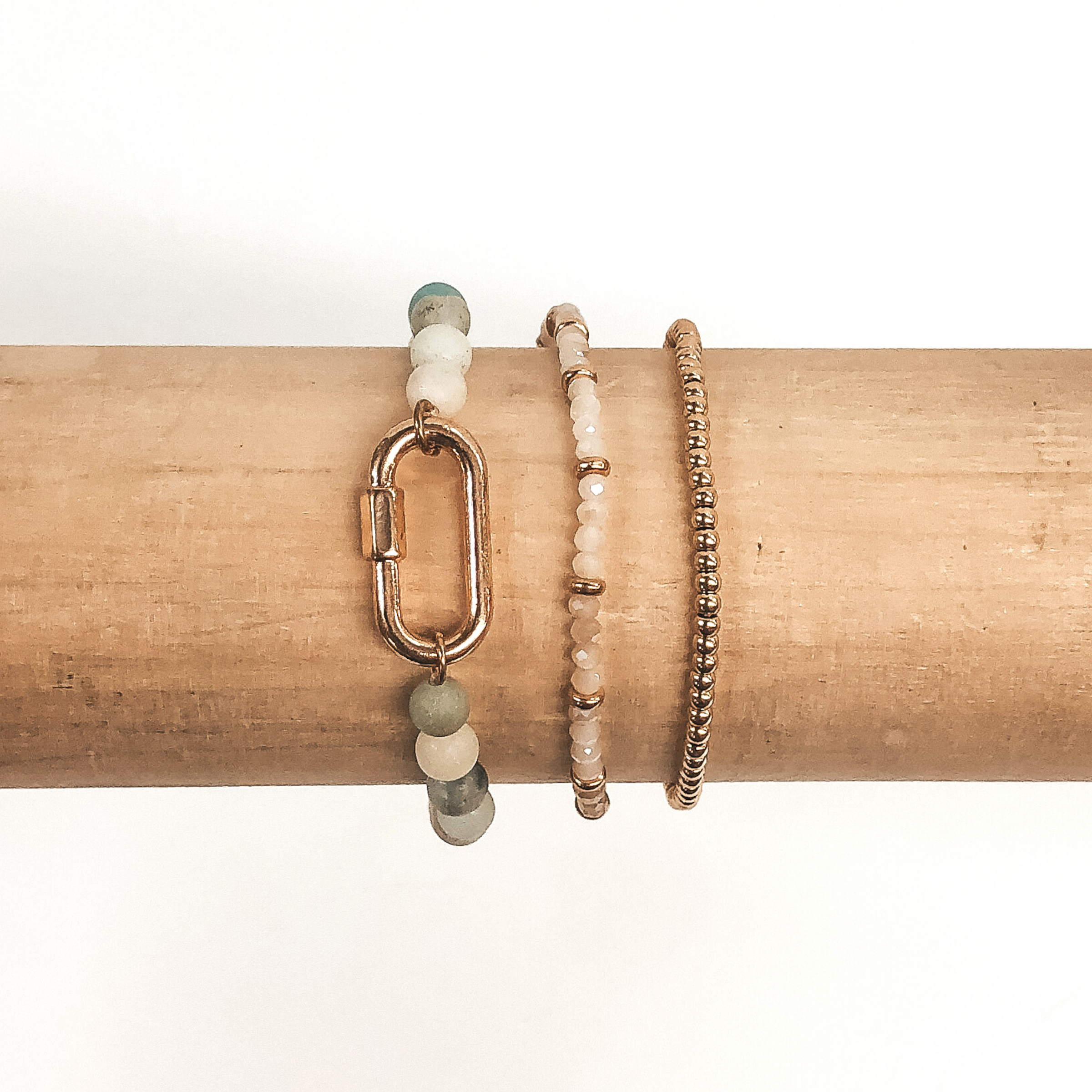 One bracelet is all gold beads, one bracelet light champagne beads and gold spacer beads, and the last bracelet has light mint marble beads with an oval pendant. These bracelets are pictured on a wood colored bracelet holder on a white background. 