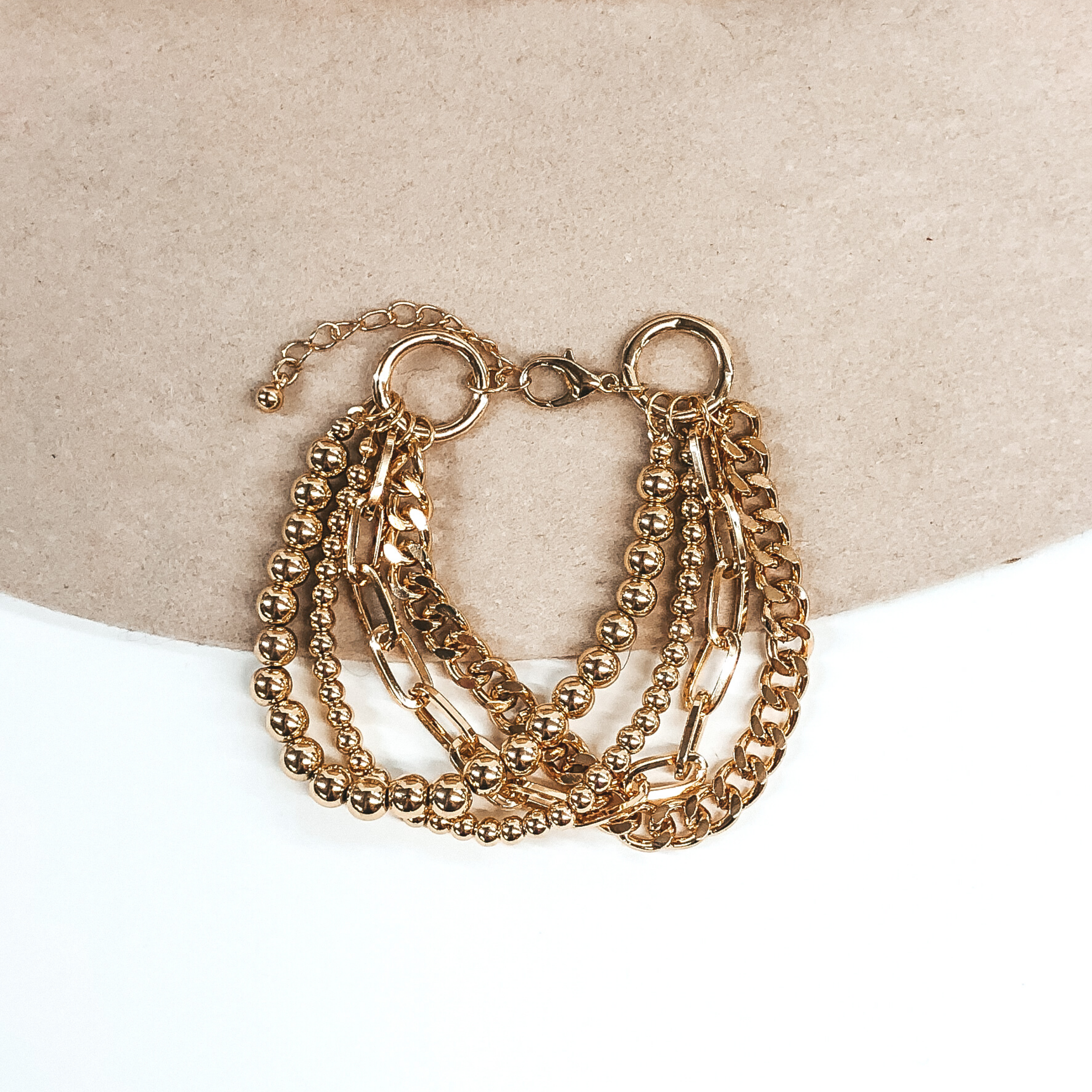 This is a gold adjustable bracelet. This bracelet includes a small and big beaded bracelet, a curb chain, and a paperclip chain. This bracelet is pictured on a beige and white background.