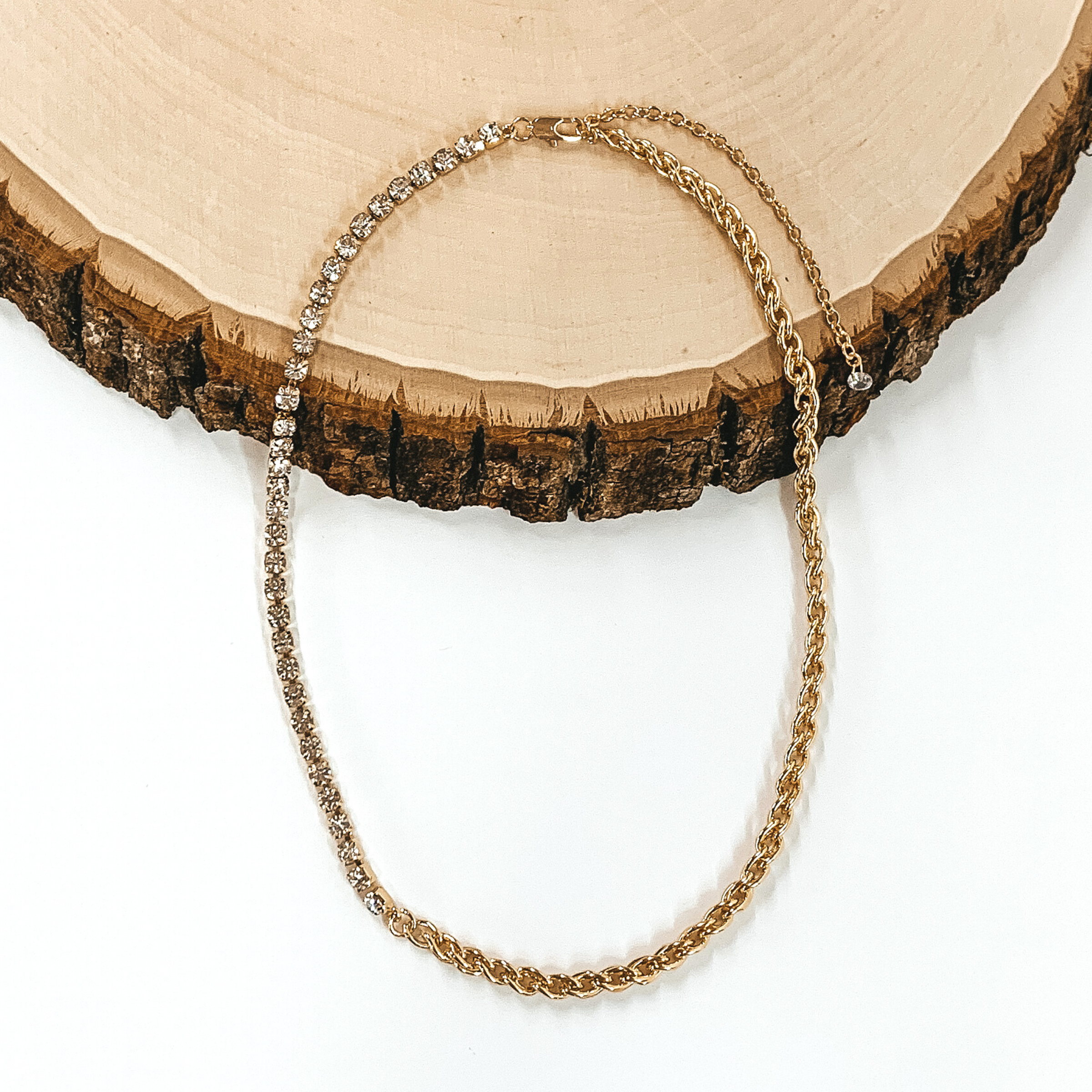 A single gold chained necklace. This necklace includes two different types of chains on the same strand. I rope strand that goes into a clear crystalized chain. This necklace is laying on a piece of wood and is pictured on a white background. 