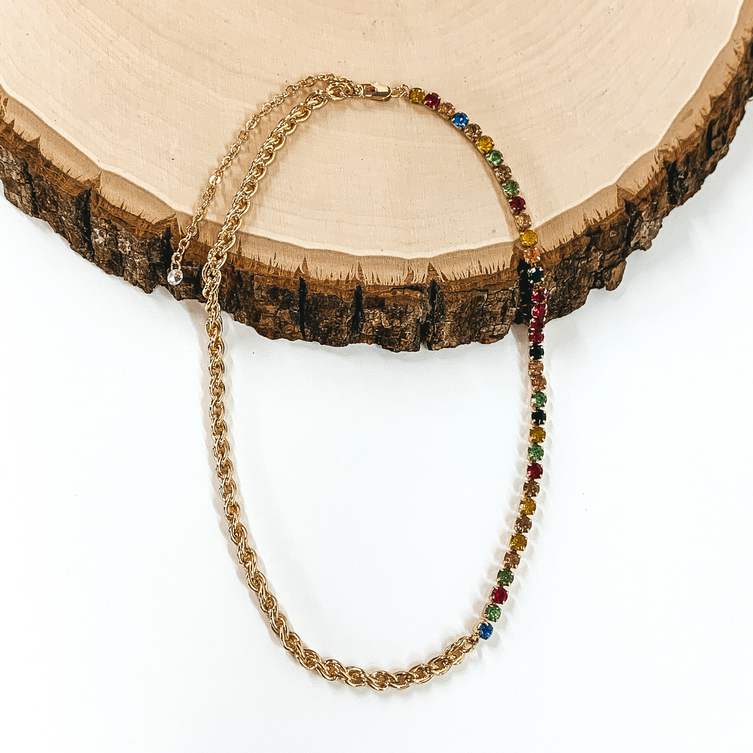 A single gold chained necklace. This necklace includes two different types of chains on the same strand. I rope strand that goes into a multicolored crystalized chain. This necklace is laying on a piece of wood and is pictured on a white background. 