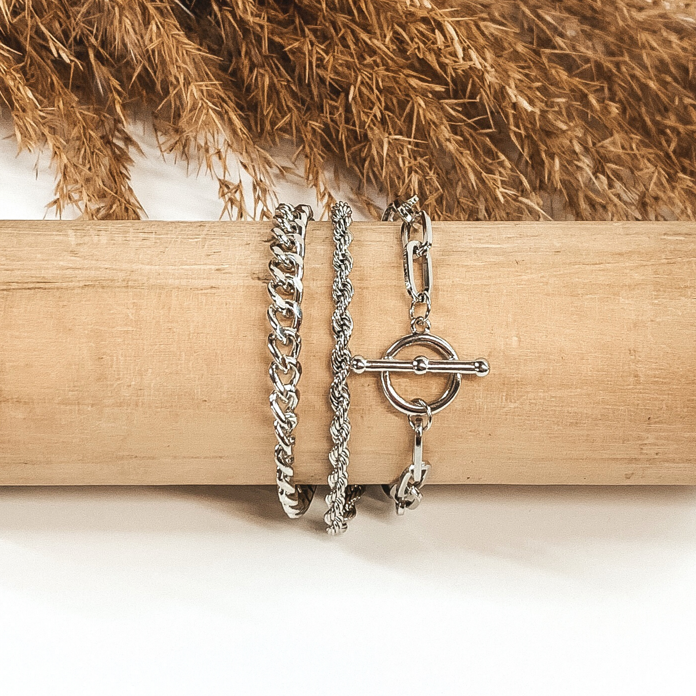 This is a three stranded silver, magnetic bracelet that includes a curb chain, a rope chain, and a paperclip chain with a toggle clasp pendant. This bracelet is pictured wrapped around a wooden bracelet holder on a white background with tan floral at the top of the picture.