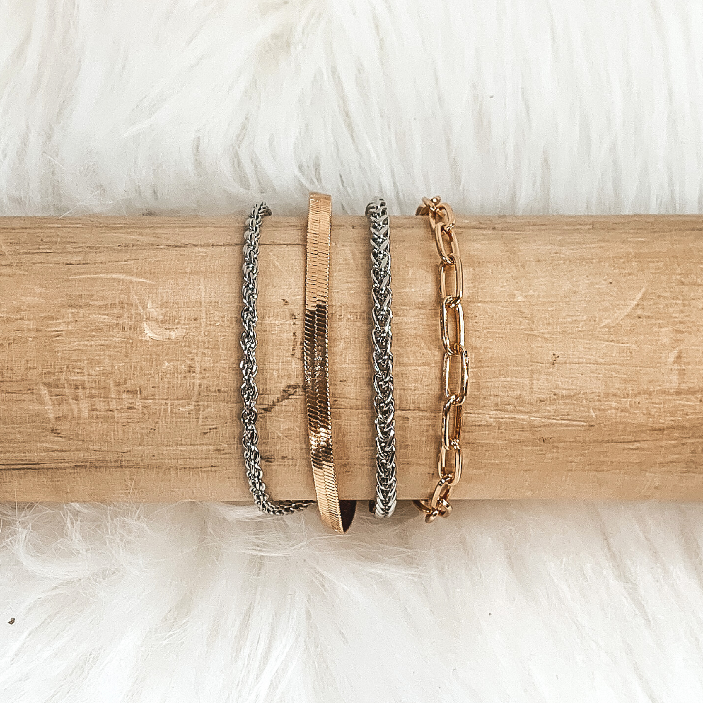 This silver and gold bracelet includes four different types of chains. This bracelet is on a light colored piece of wood pictured on a white fur background. 