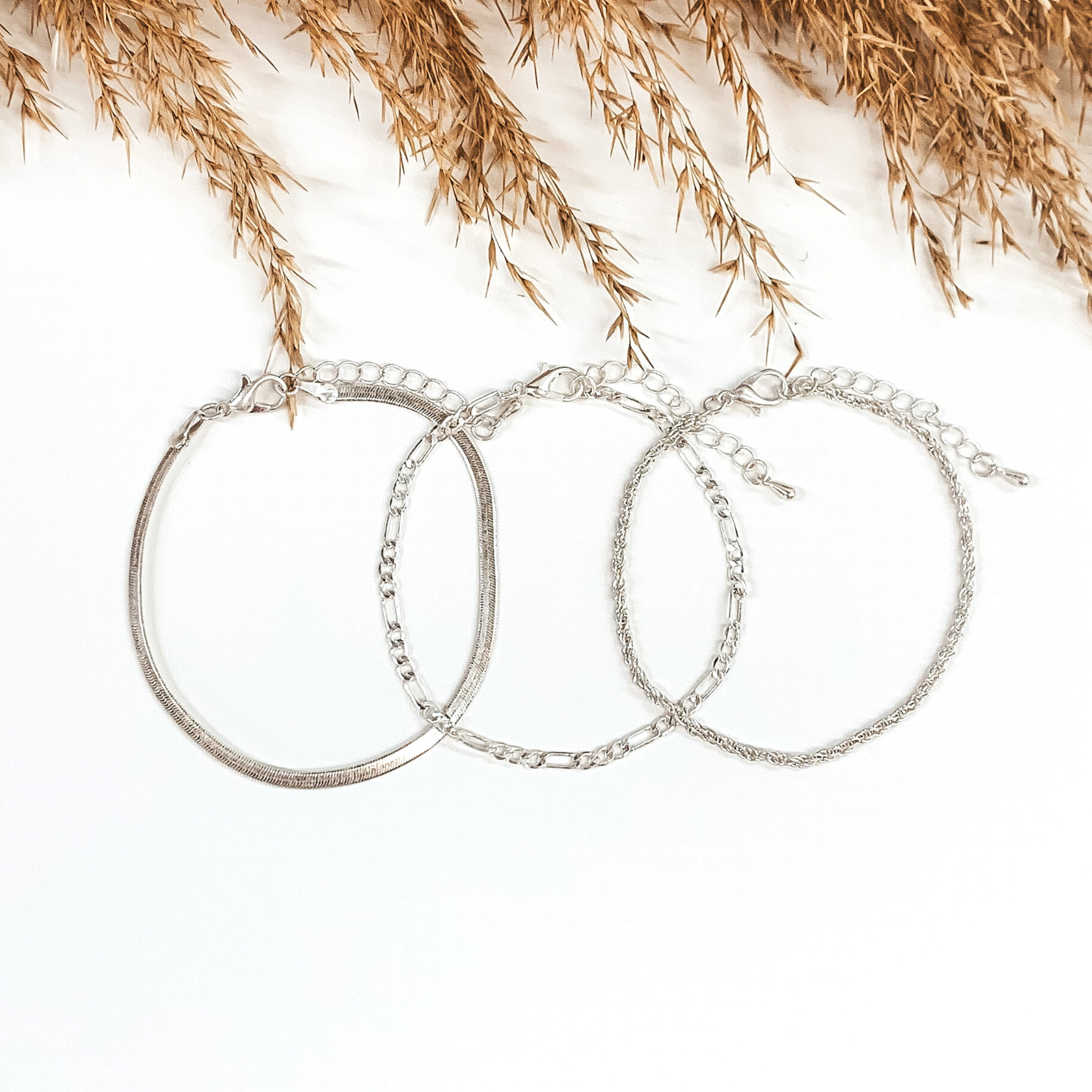 Three silver chained bracelets that are laying on top of each other. There is a snake chain, figaro chain, and a rope chain bracelet. These bracelets are pictured on a white background with tan floral at the top of the picture. 